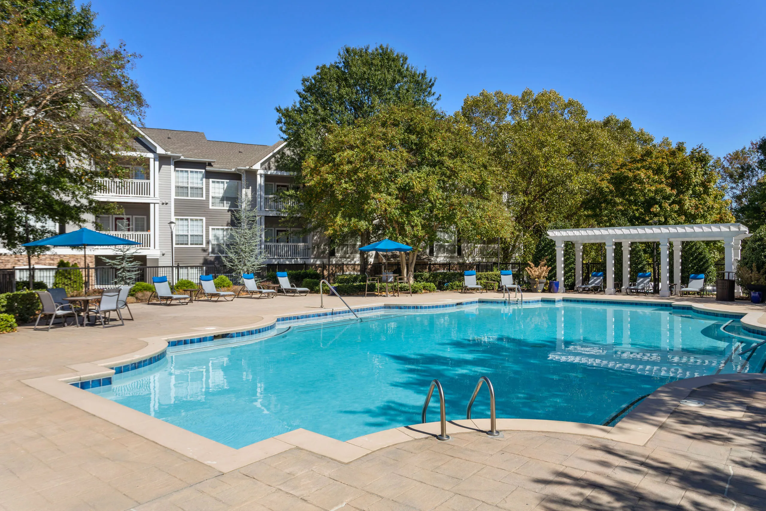 Elegant apartment complex with a sparkling swimming pool, surrounded by lush greenery and stylish outdoor seating under blue umbrellas.