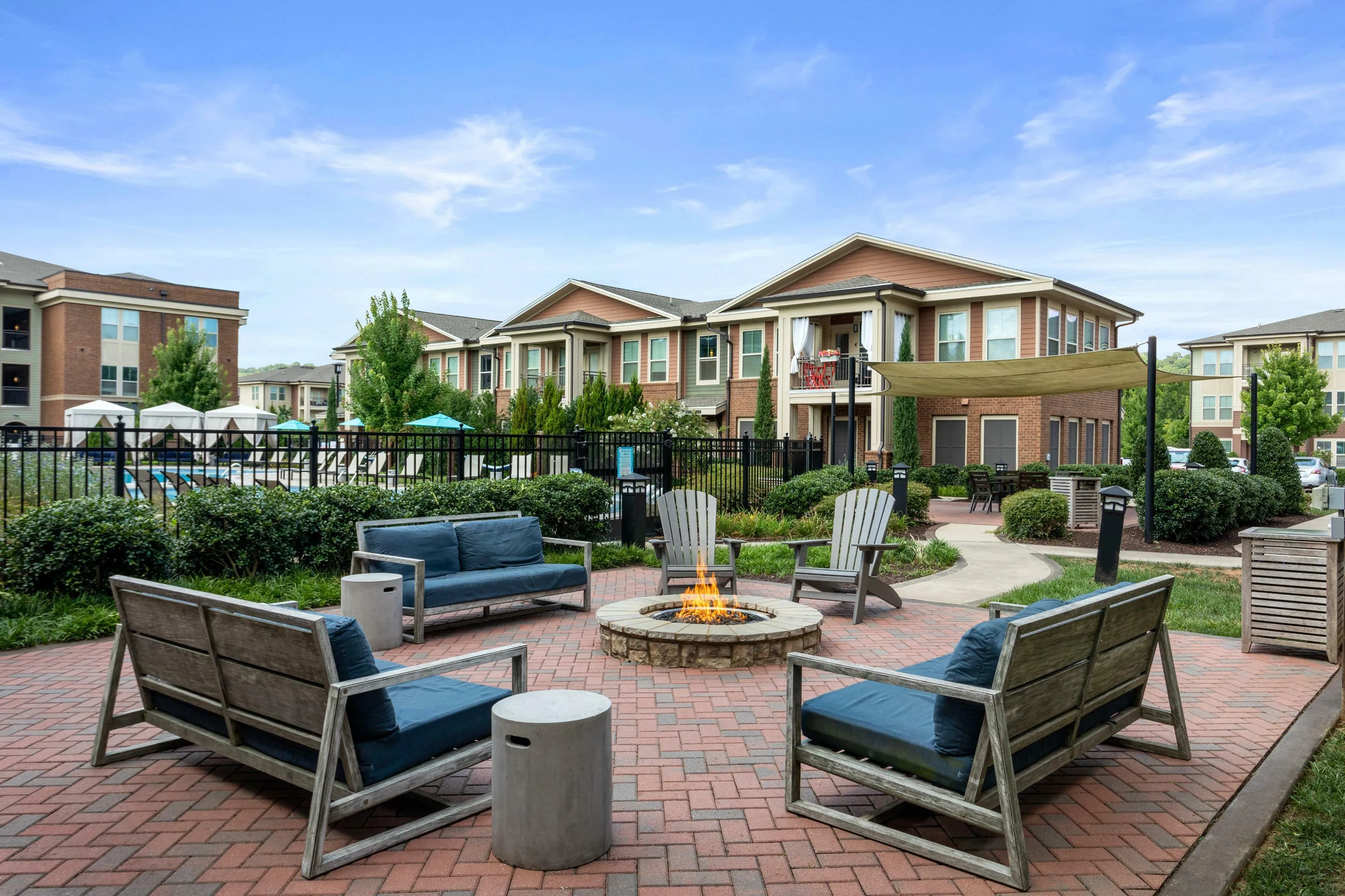 Inviting outdoor seating area with fire pit surrounded by greenery and modern apartment buildings under a clear blue sky.