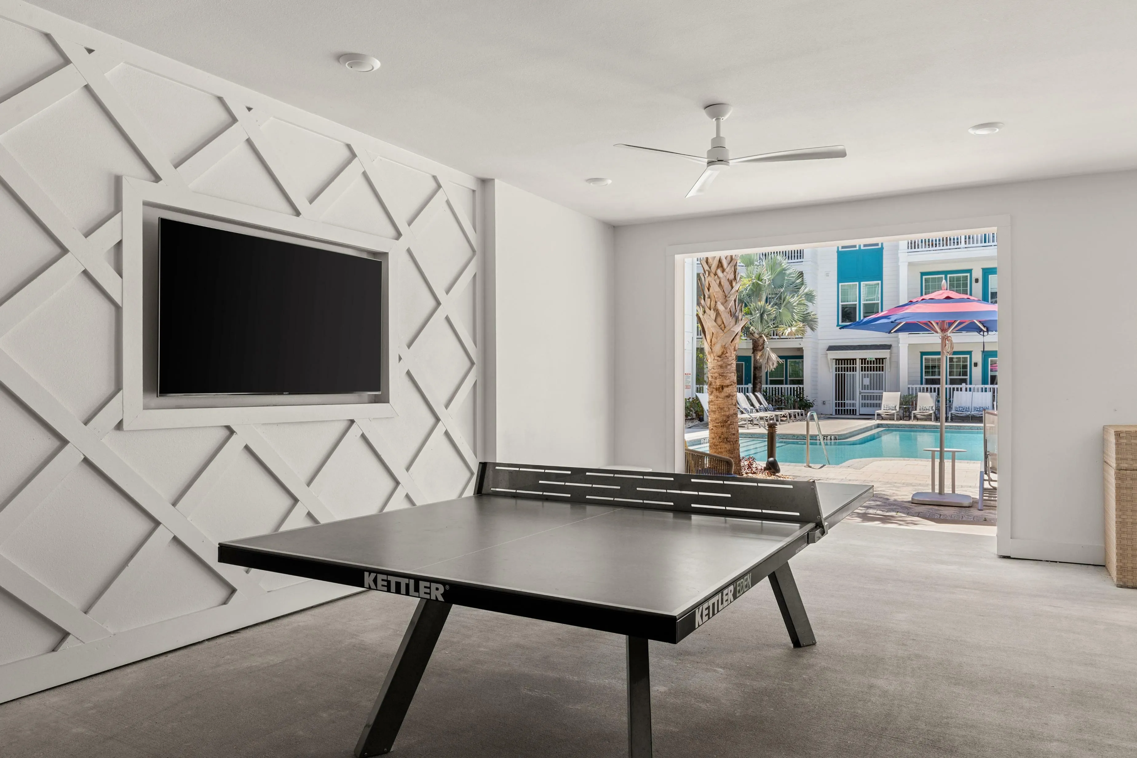 Luxurious recreation room featuring a stylish ping pong table, modern wall design, and a stunning view of the sunlit pool area with palm trees and umbrellas.