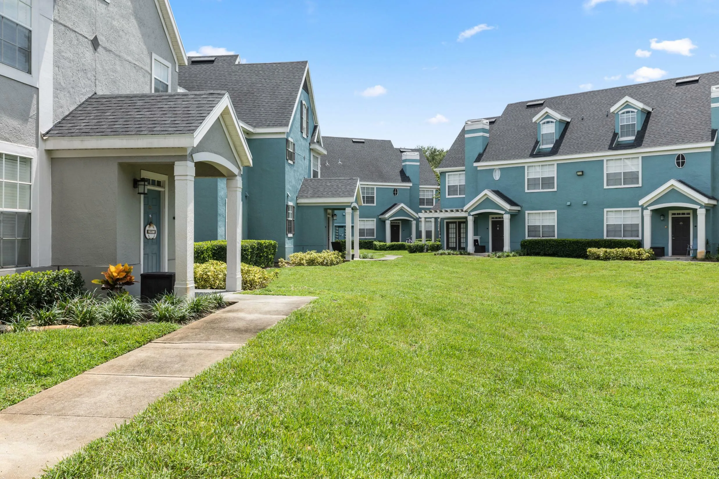 Charming townhouse community with well-manicured lawns, classic architectural details, and inviting blue and white facades under a clear blue sky.