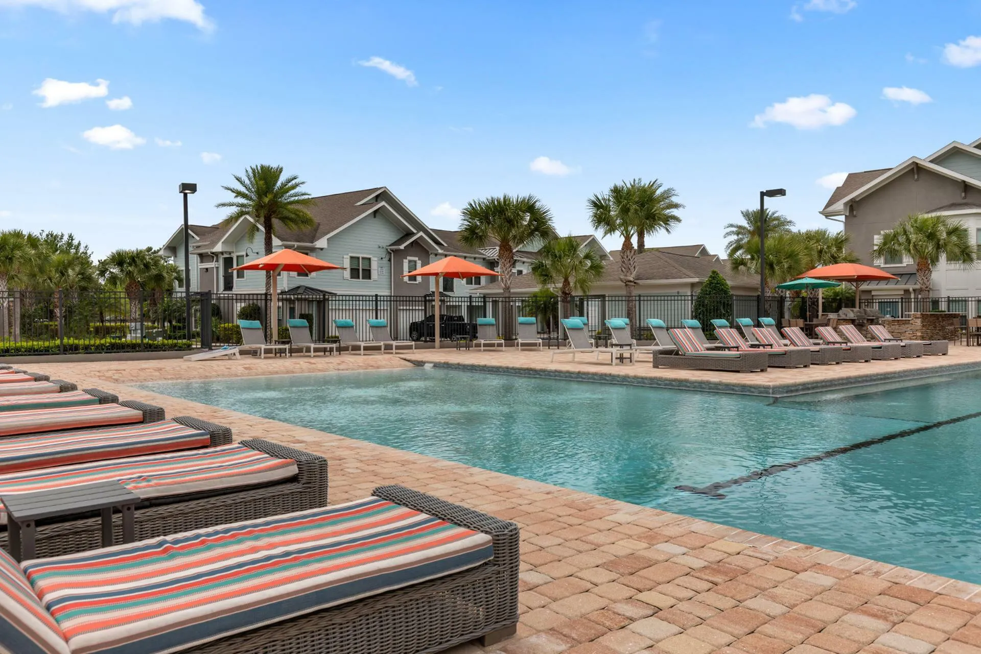 Luxurious poolside view at a modern apartment complex with relaxing sun loungers, colorful umbrellas, and vibrant palm trees under a bright blue sky.