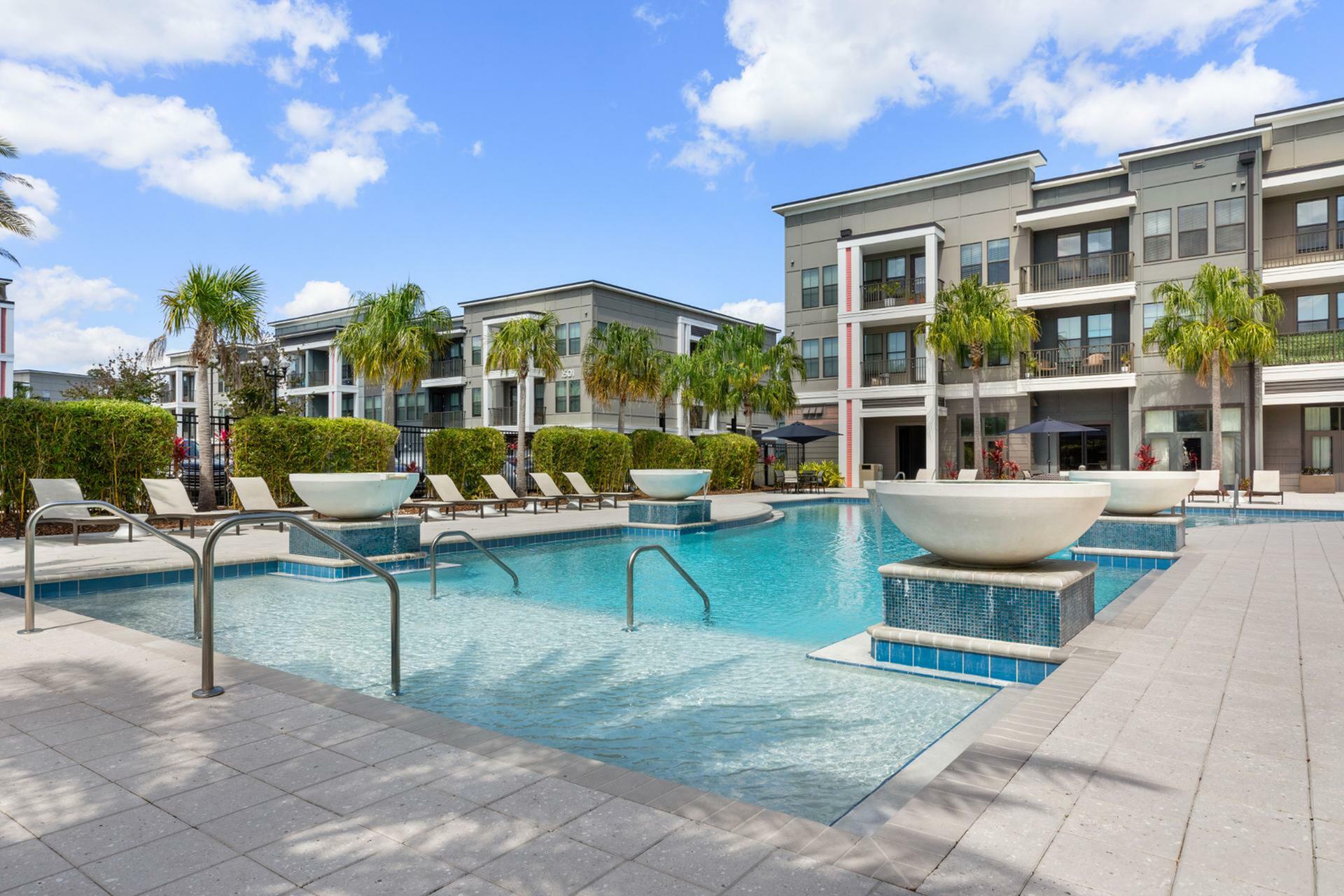 Luxurious apartment complex featuring a sparkling pool, chic loungers, and lush palm trees, set against a clear blue sky.