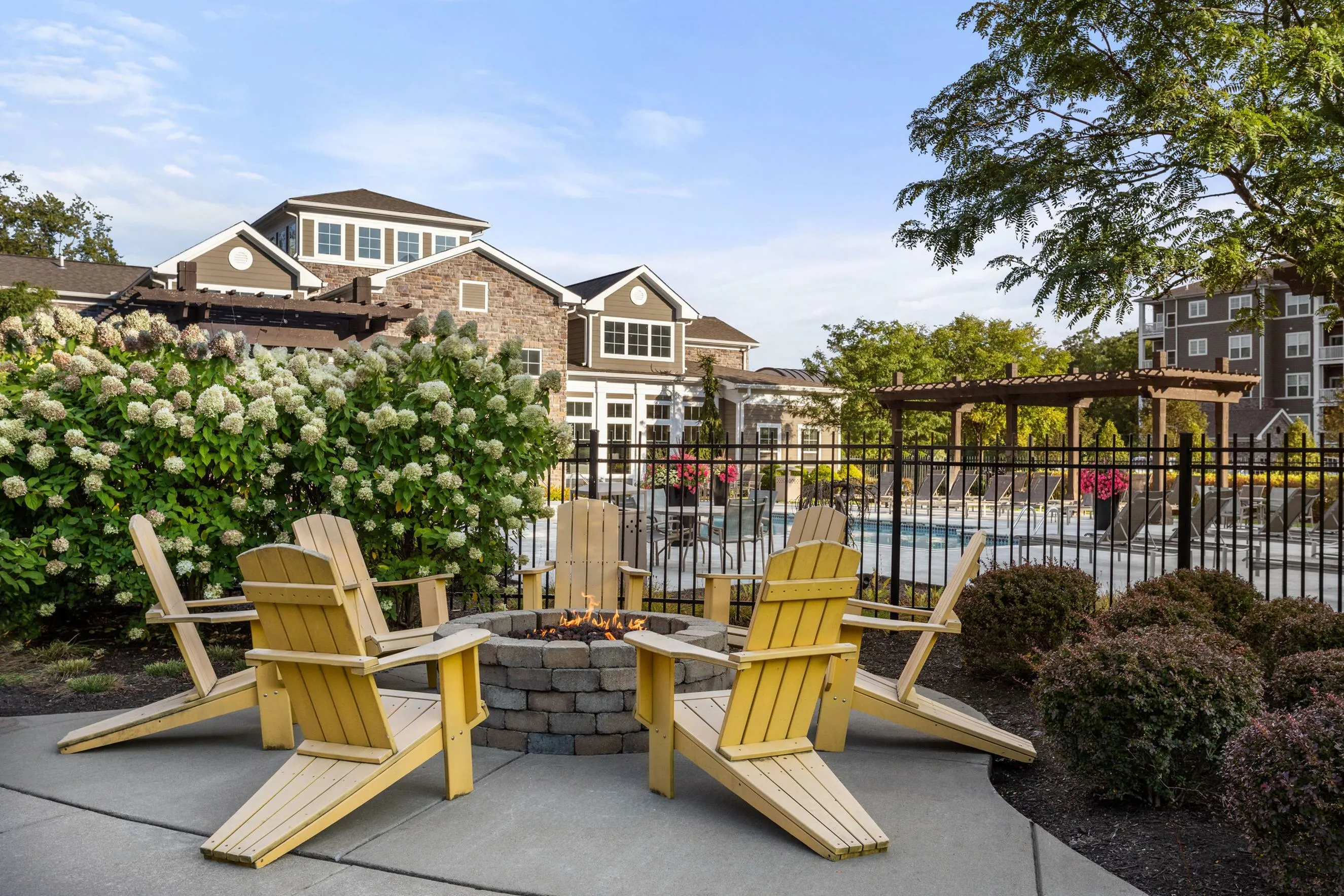 Charming outdoor setting with cozy fire pit surrounded by Adirondack chairs, lush greenery, and a refreshing pool in the background.
