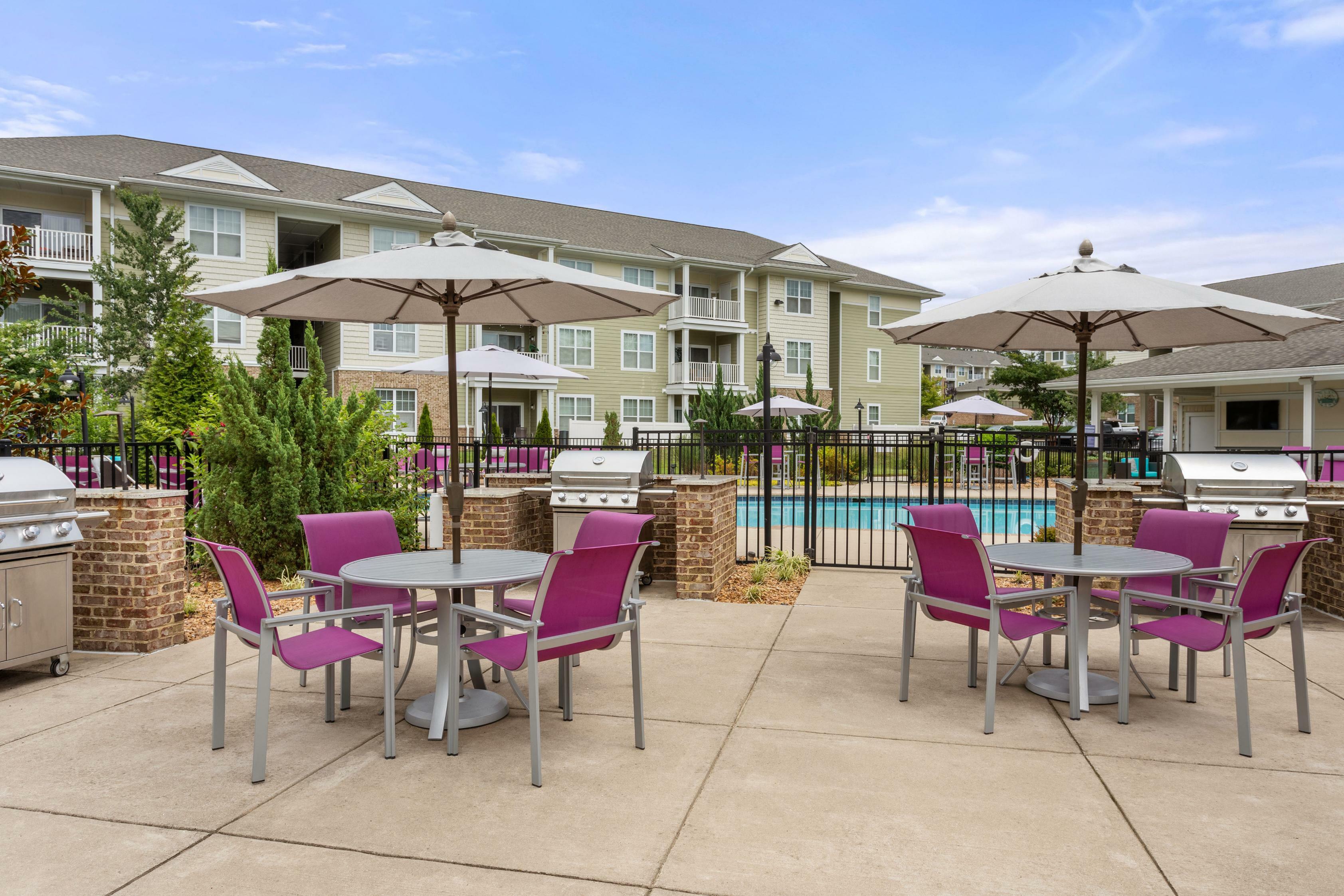 Charming apartment patio area featuring vibrant purple chairs, inviting parasols, and modern barbecue grills, with lush greenery and a sparkling pool in the background.