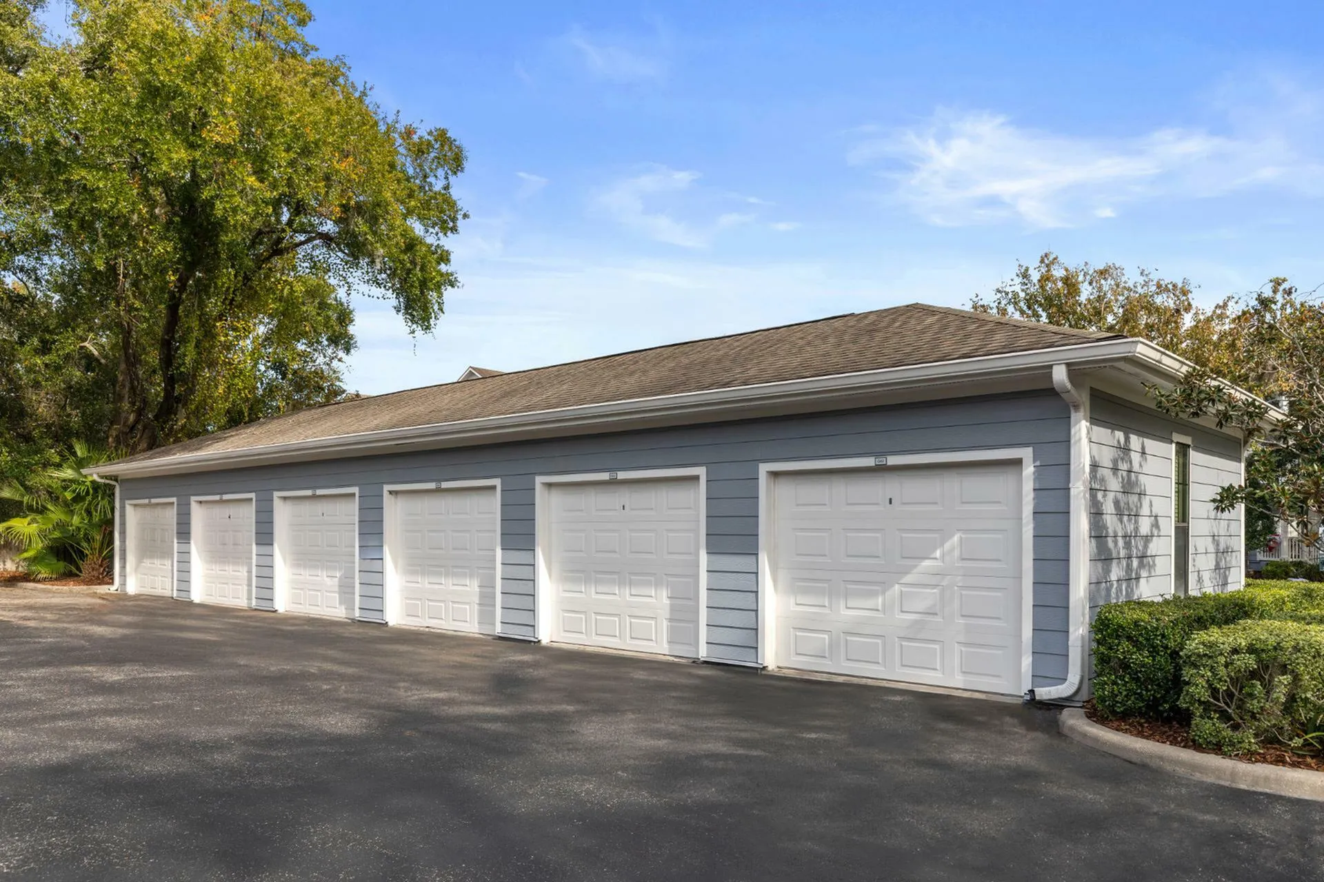Charming residential garage structure with multiple white doors, set against lush greenery and a clear blue sky.