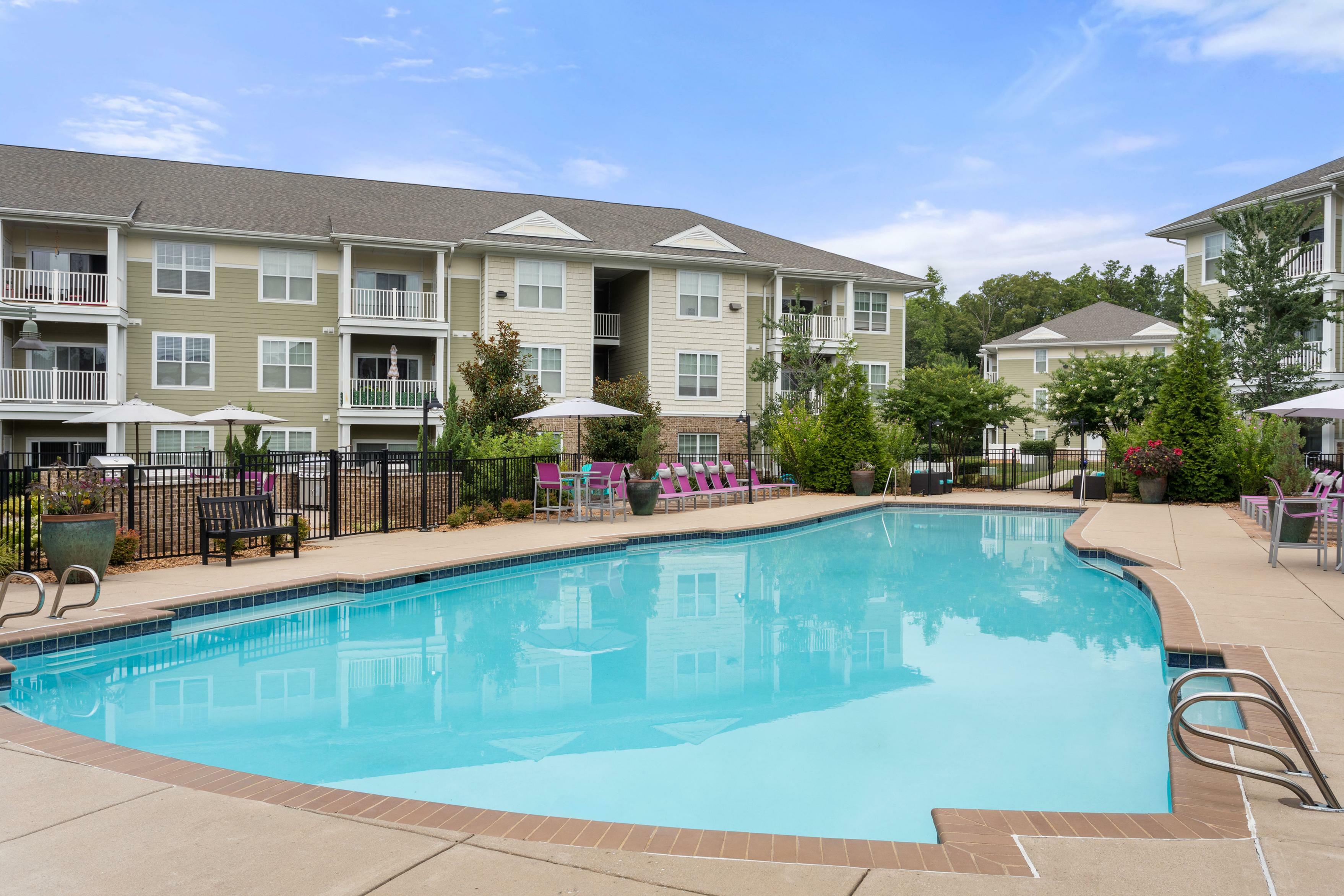 Charming apartment complex featuring a bright blue swimming pool, surrounded by manicured greenery and outdoor seating for a relaxing ambience.