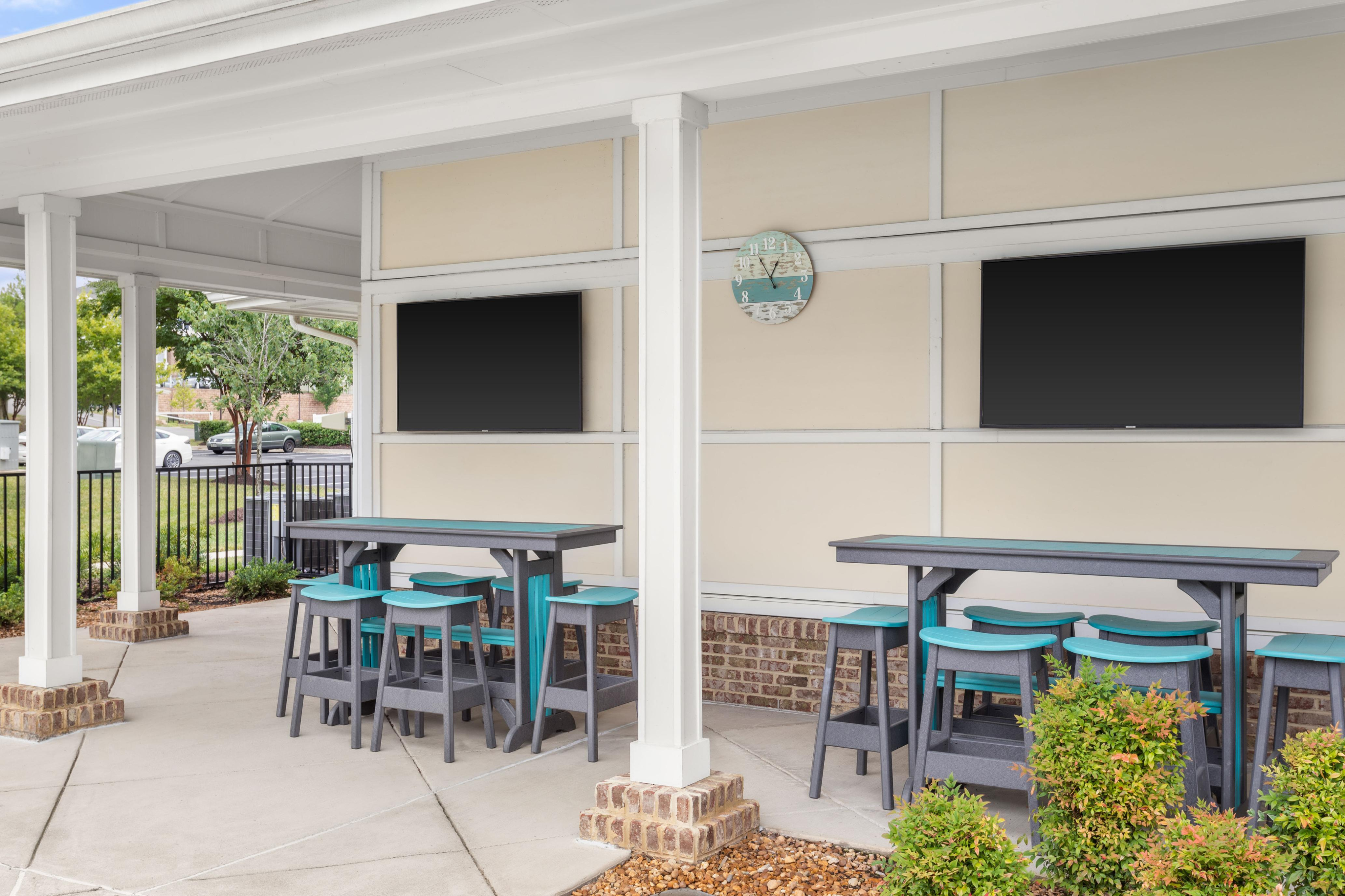 Inviting outdoor lounge area with modern bar stools and tables, framed by lush greenery and equipped with dual flat-screen TVs for entertainment.