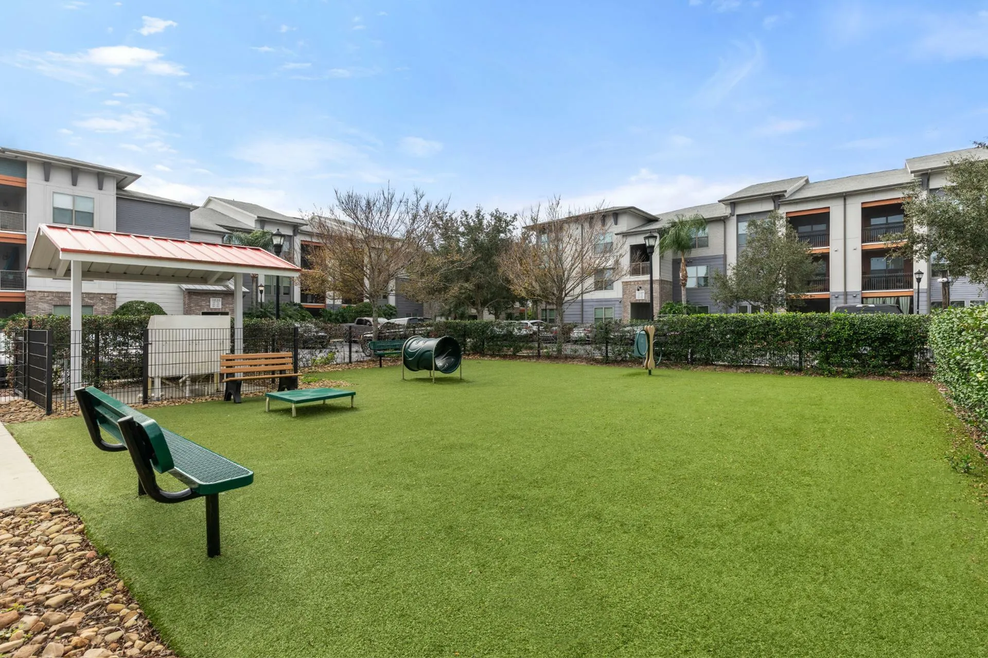 Inviting pet-friendly outdoor area with benches, agility equipment, and lush green space surrounded by modern apartment buildings.