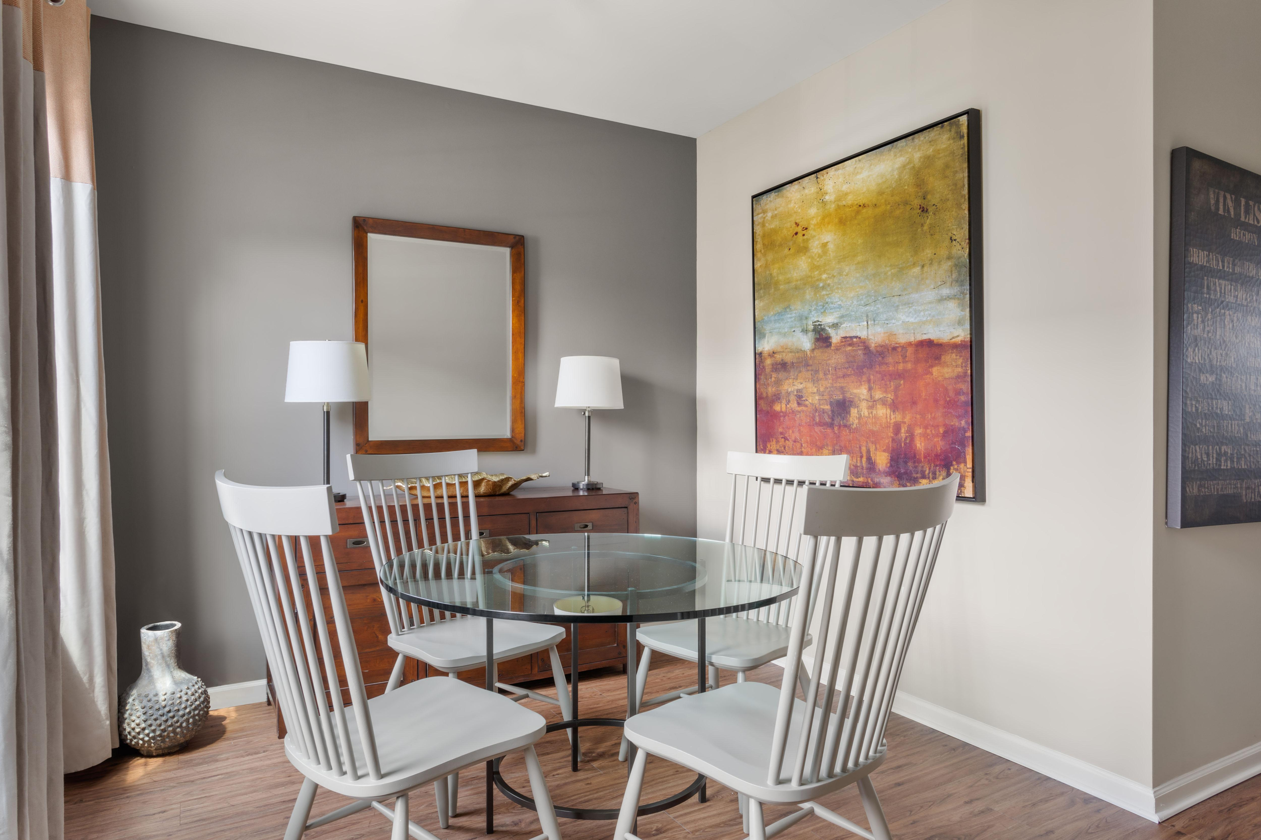 Modern dining area with a sleek glass round table, surrounded by elegant white chairs. Features vibrant abstract artwork and a warm wooden sideboard, creating a contemporary and inviting atmosphere.