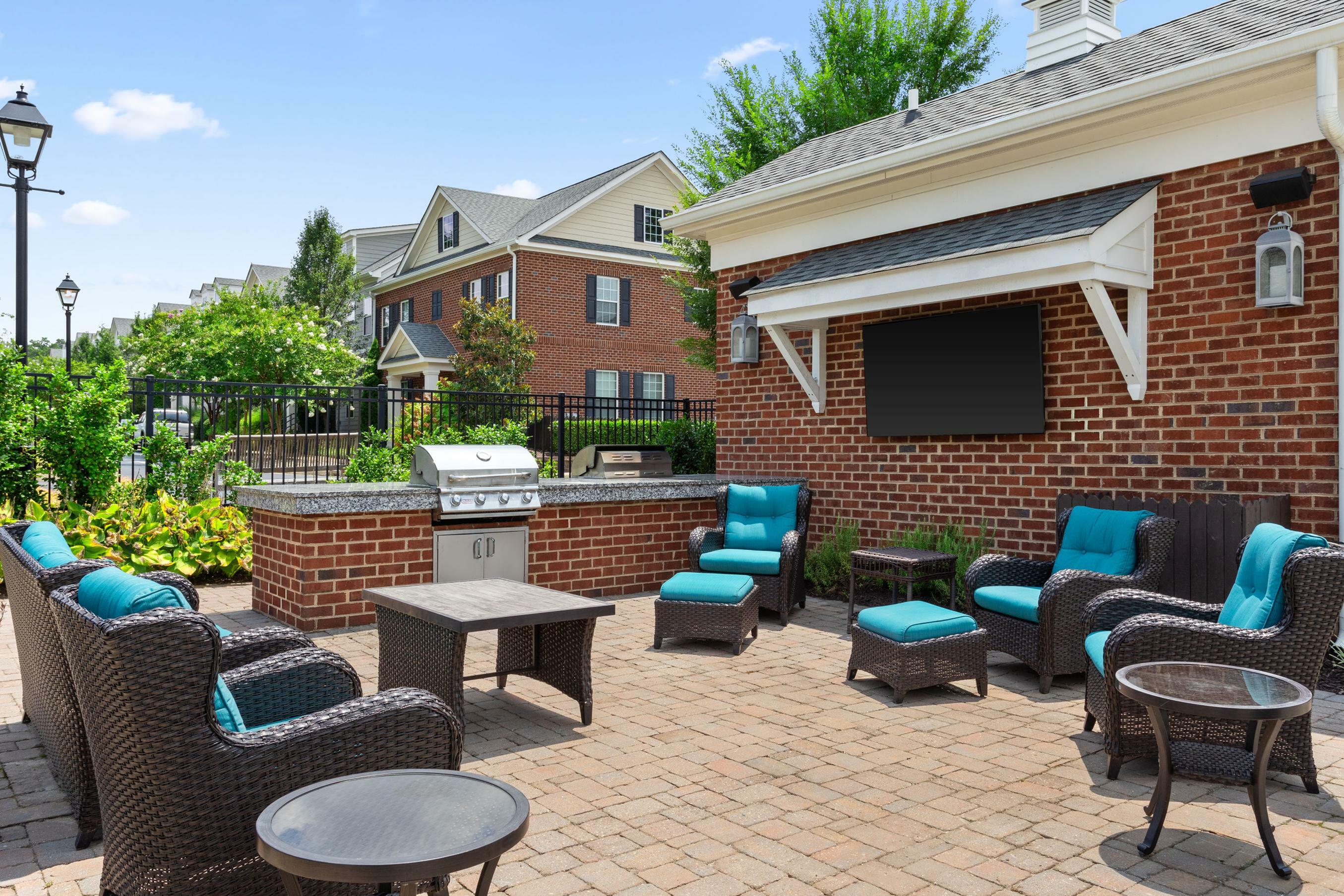 Charming outdoor patio with comfortable seating, vibrant blue cushions, and a sleek barbecue grill surrounded by lush greenery and classic brick architecture.