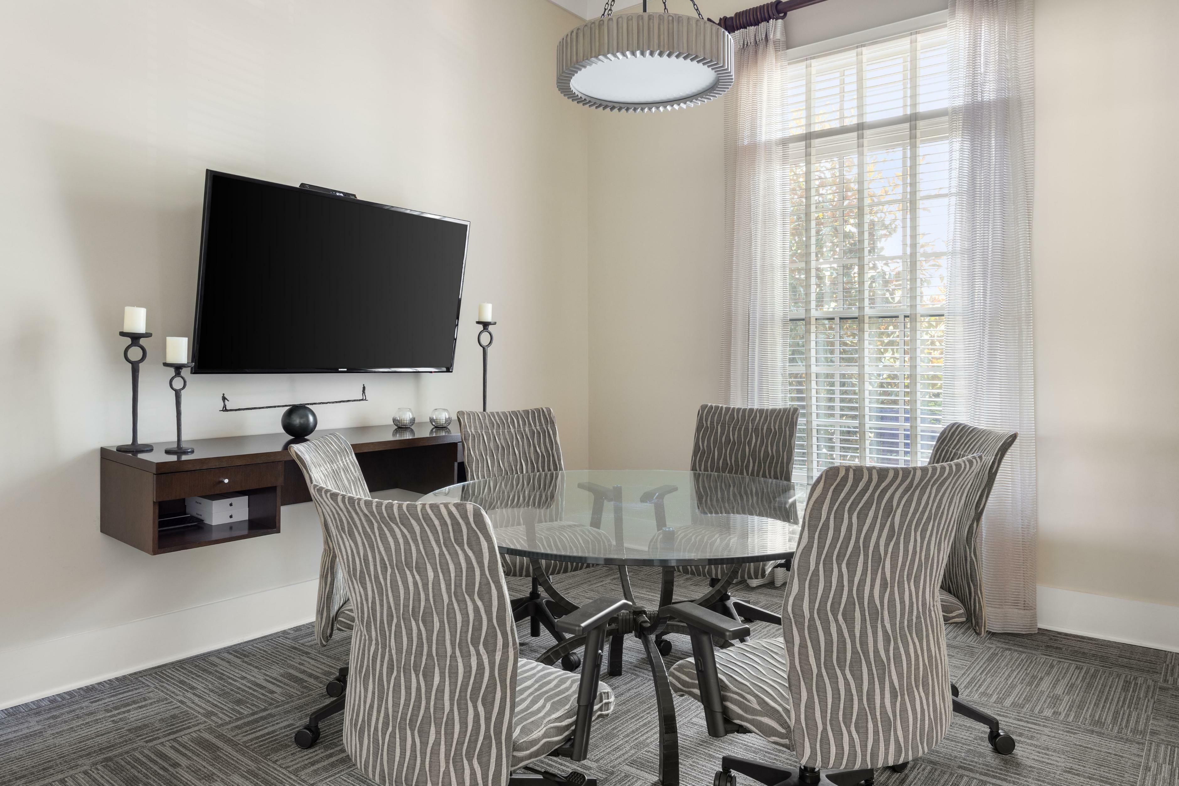 Elegant meeting room with a modern glass table, six stylish chairs, and a wall-mounted TV, flooded with natural light from large windows.