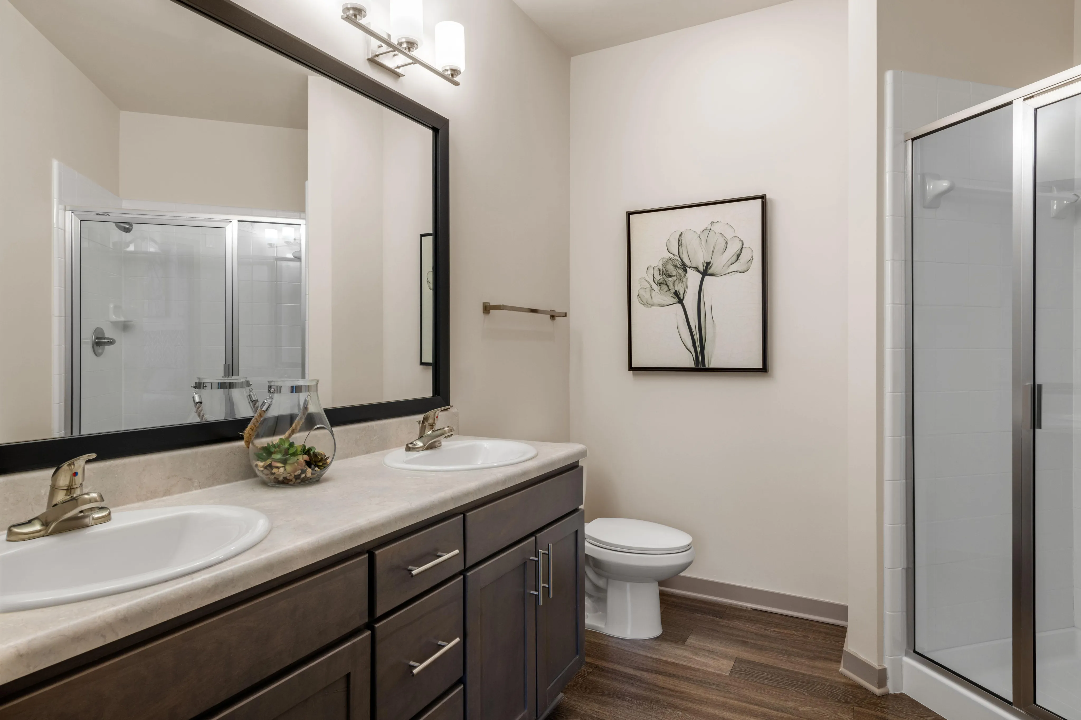 Elegant bathroom design featuring dual sinks with modern fixtures, stylish dark wood cabinetry, framed floral artwork, and a spacious glass-enclosed shower.