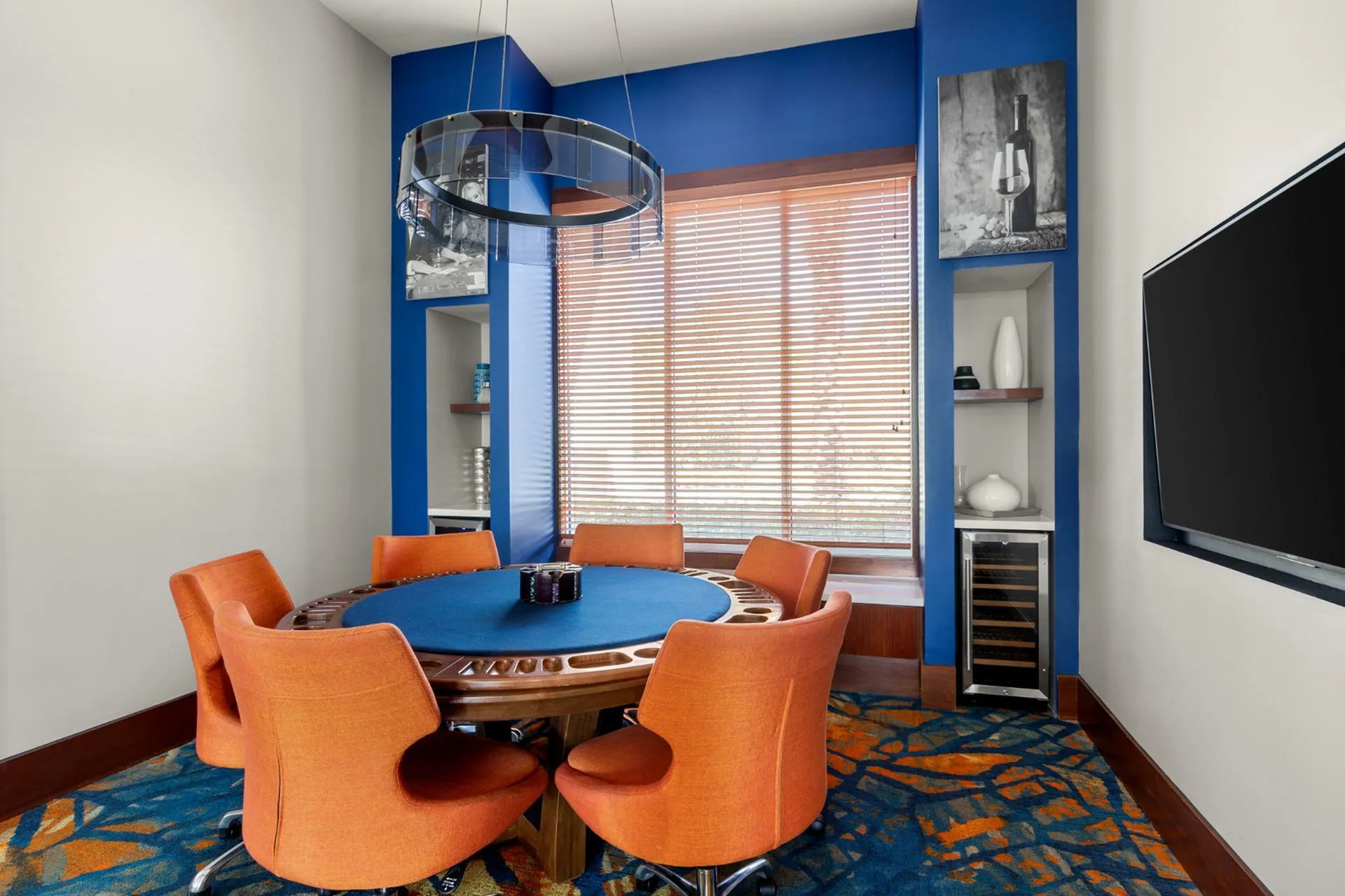 Vibrant entertainment room with a stylish poker table surrounded by bold orange chairs, complemented by sleek blue walls and modern decor.