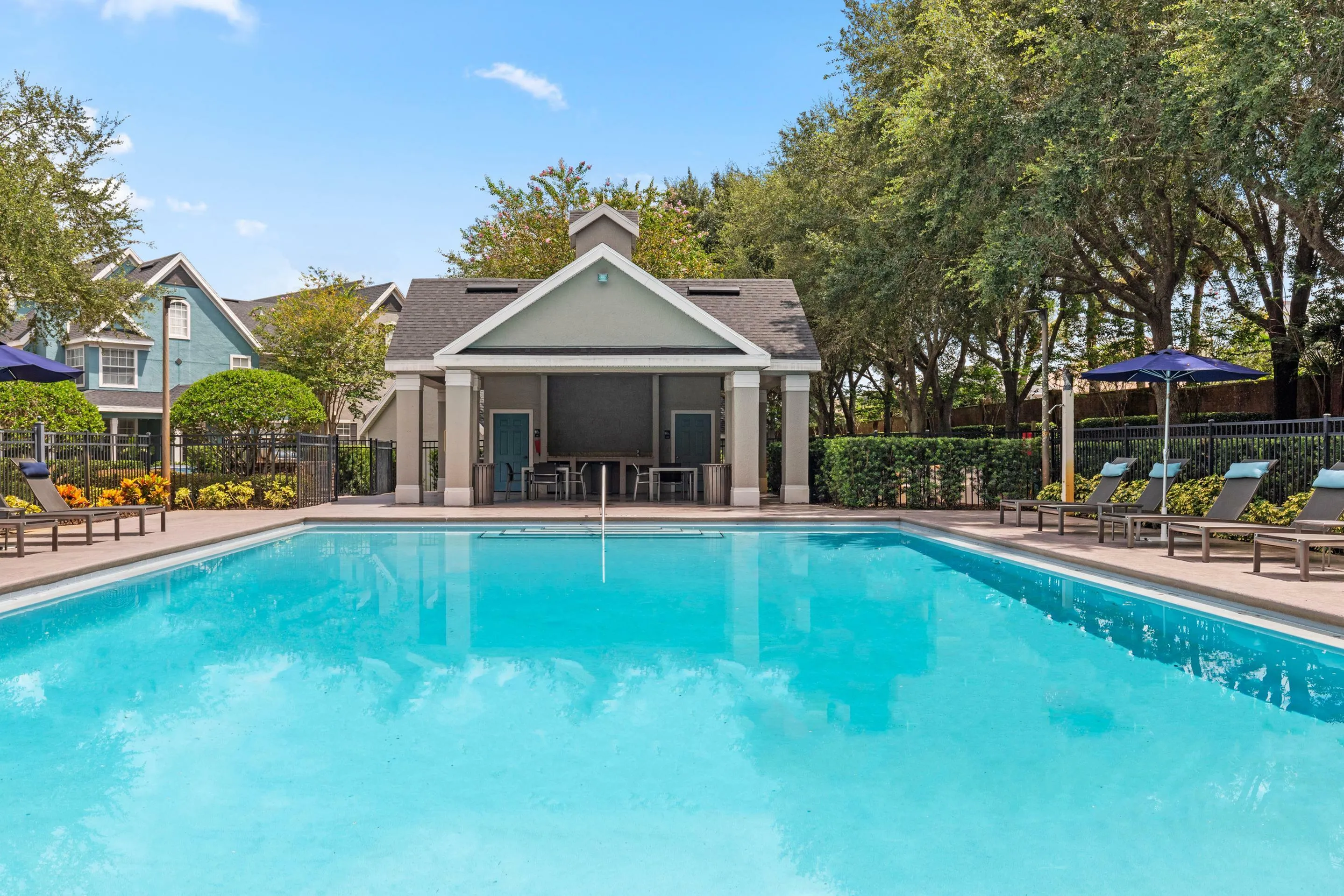 Inviting outdoor pool area with elegant lounge chairs and shaded cabanas, surrounded by lush greenery and charming residential architecture.
