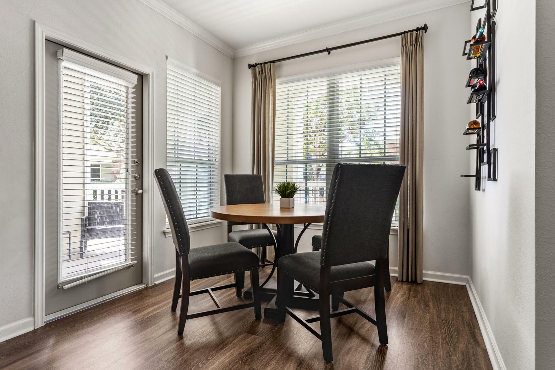 Bright and cozy dining nook with a round wooden table and elegant upholstered chairs, featuring large windows with blinds and a view of the greenery outside.