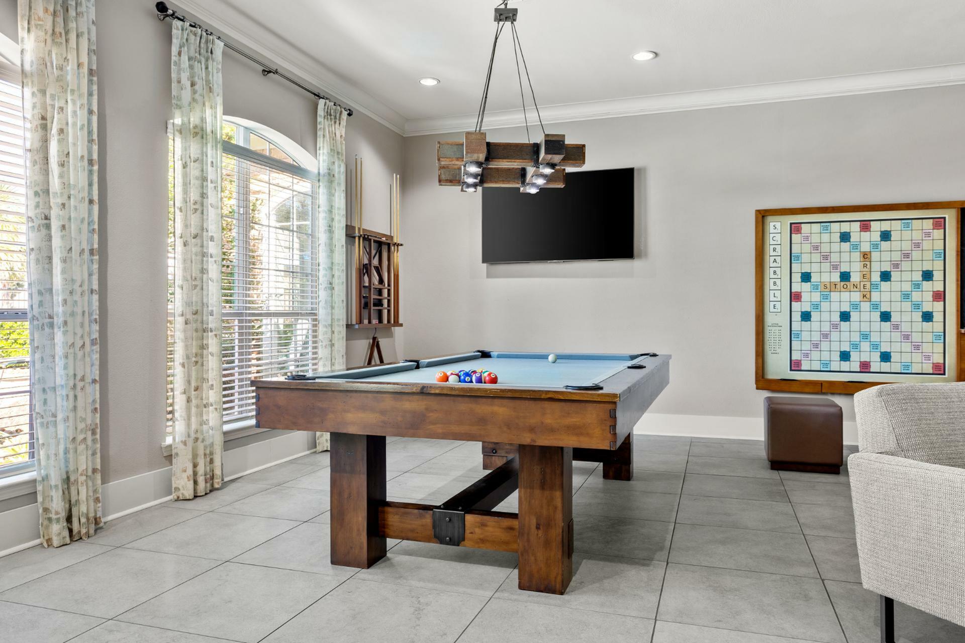 Elegant game room featuring a classic pool table under stylish lighting, with a decorative wall-mounted Scrabble board and large windows inviting natural light.