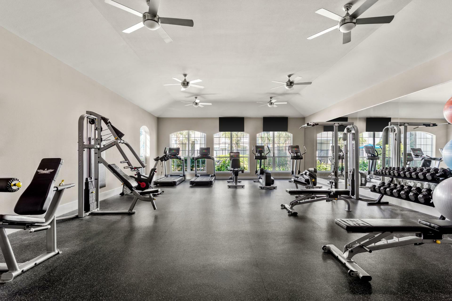 Luxurious apartment gym with modern equipment, including treadmills, weight machines, and free weights, featuring natural light and ceiling fans for a refreshing workout experience.
