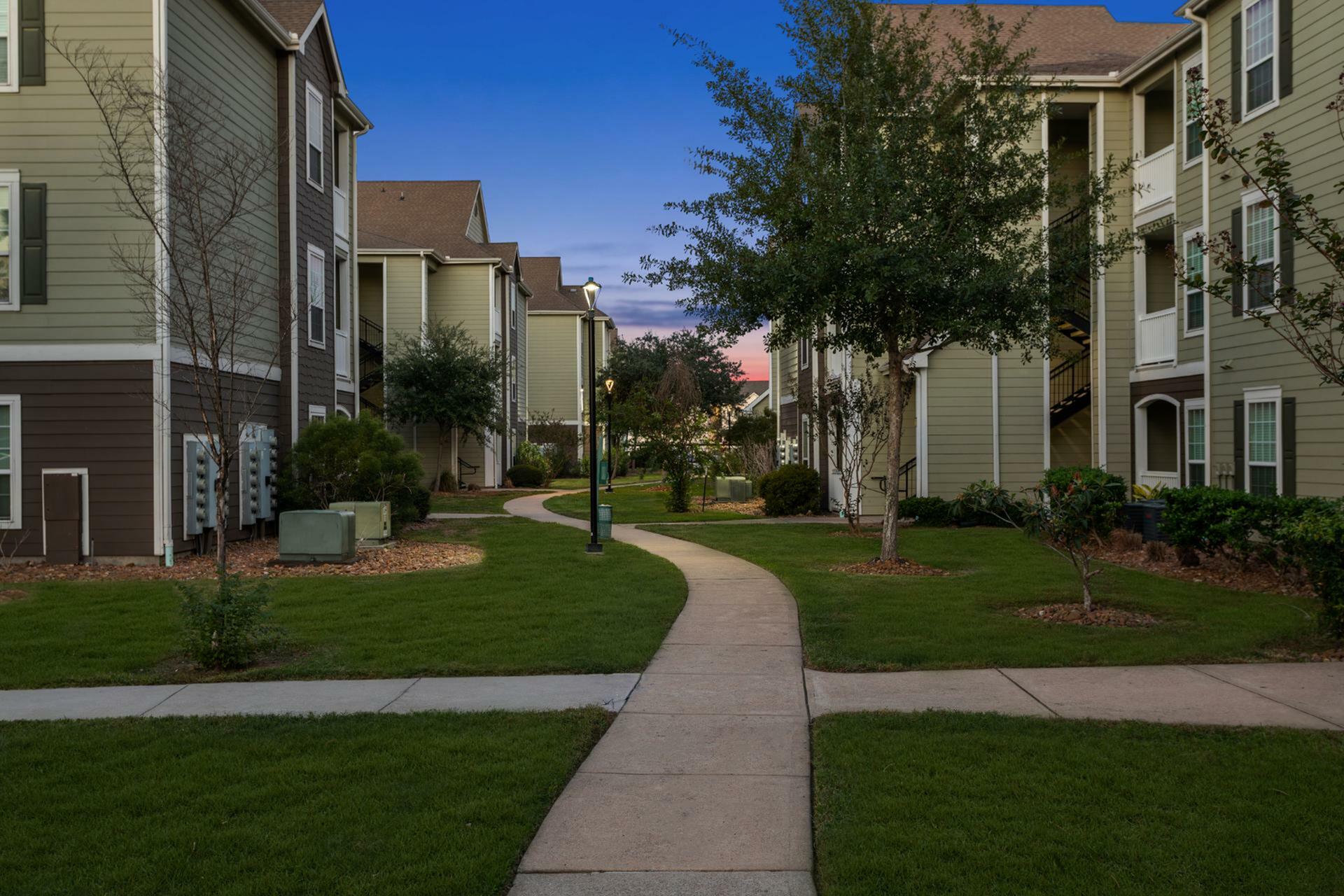 Charming suburban apartment complex with scenic pathways, lush greenery, and inviting outdoor spaces at sunset.