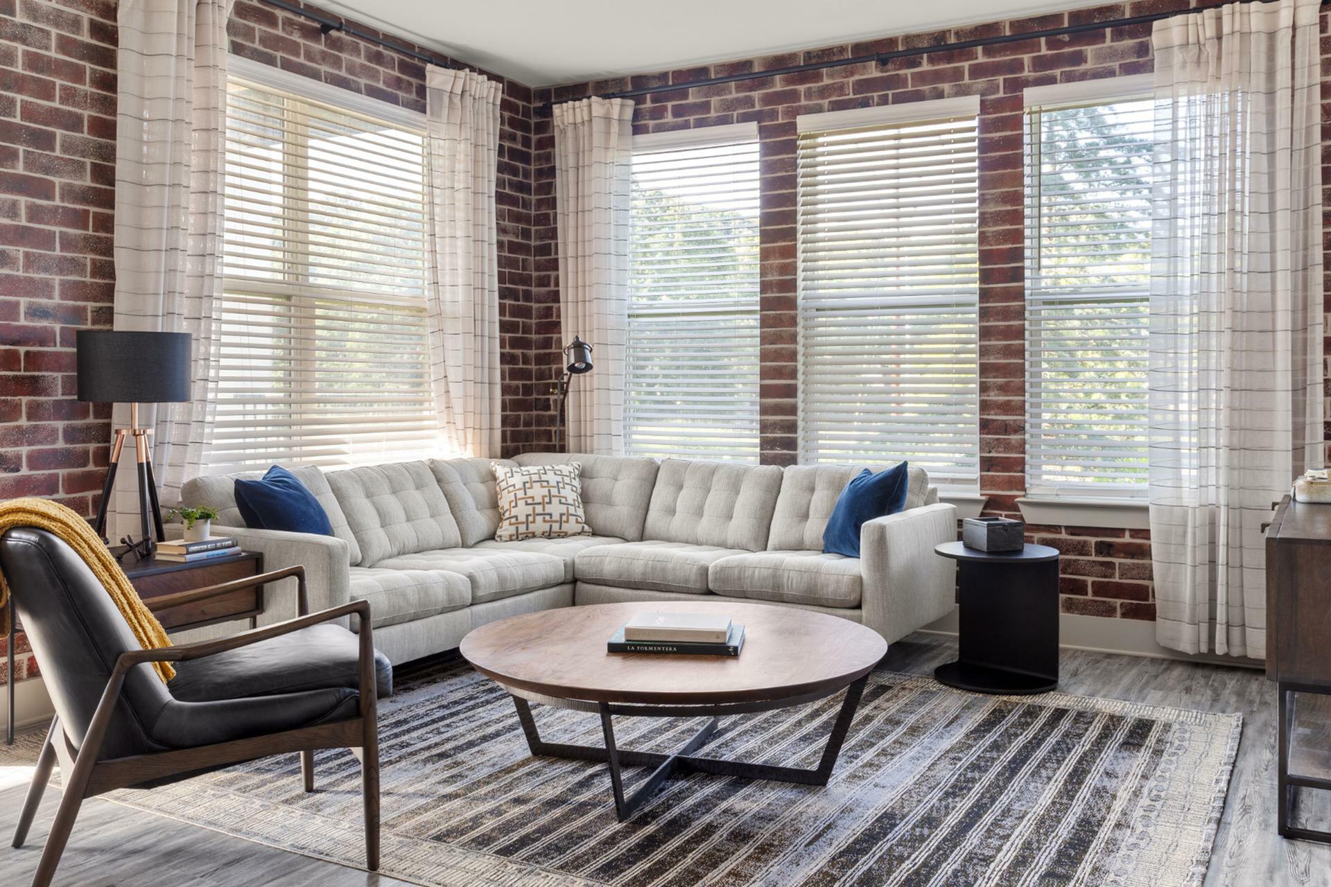 Cozy living room with modern decor, featuring a plush sectional sofa, stylish accent chair, and natural light streaming through large windows with brick walls.
