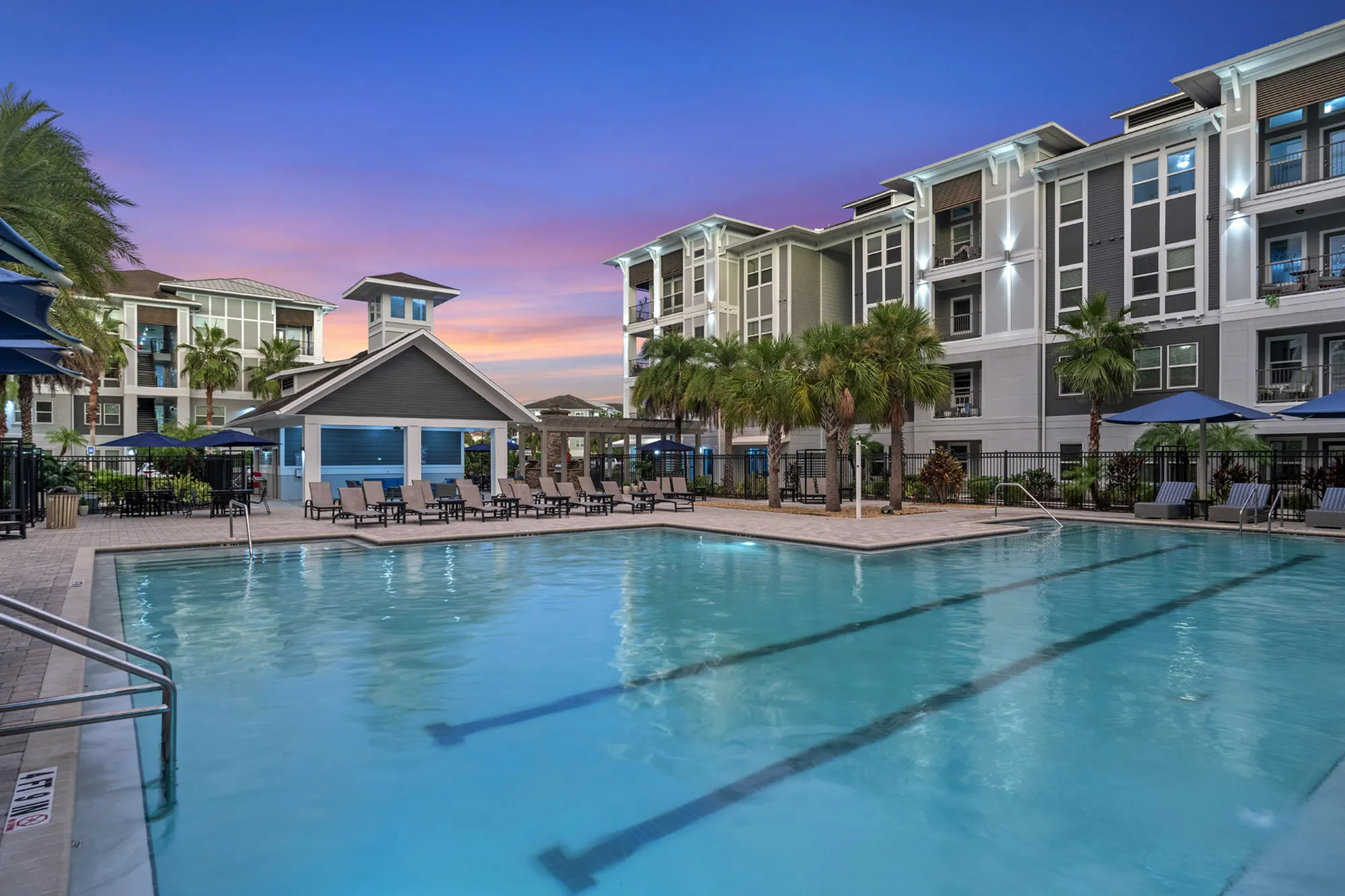 Luxurious apartment complex featuring a pristine swimming pool, surrounded by palm trees and elegant lounge chairs, set against a stunning sunset backdrop.