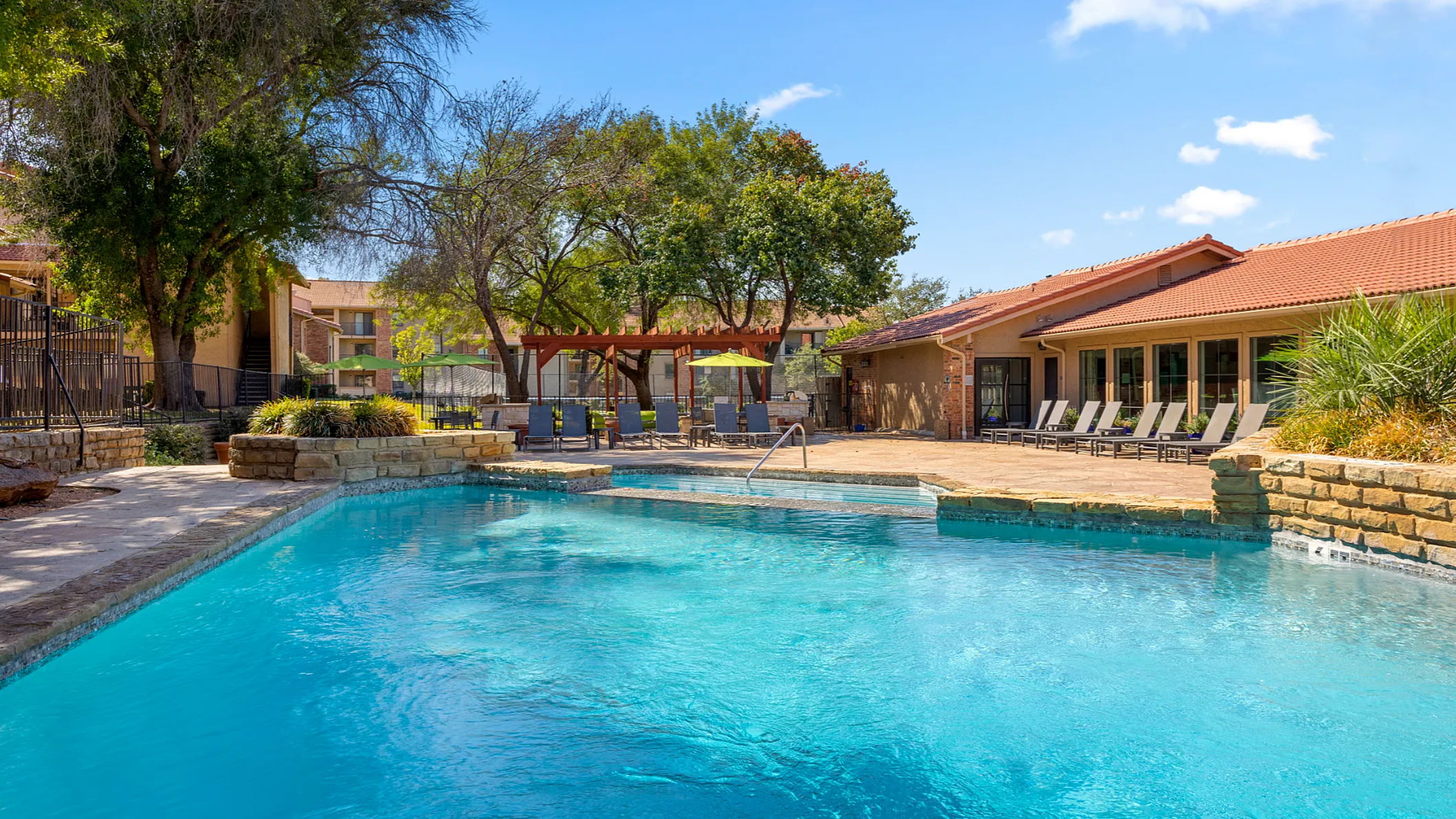 Inviting apartment complex pool area with crystal-clear water, surrounded by lush greenery and a charming sun patio with lounge chairs.