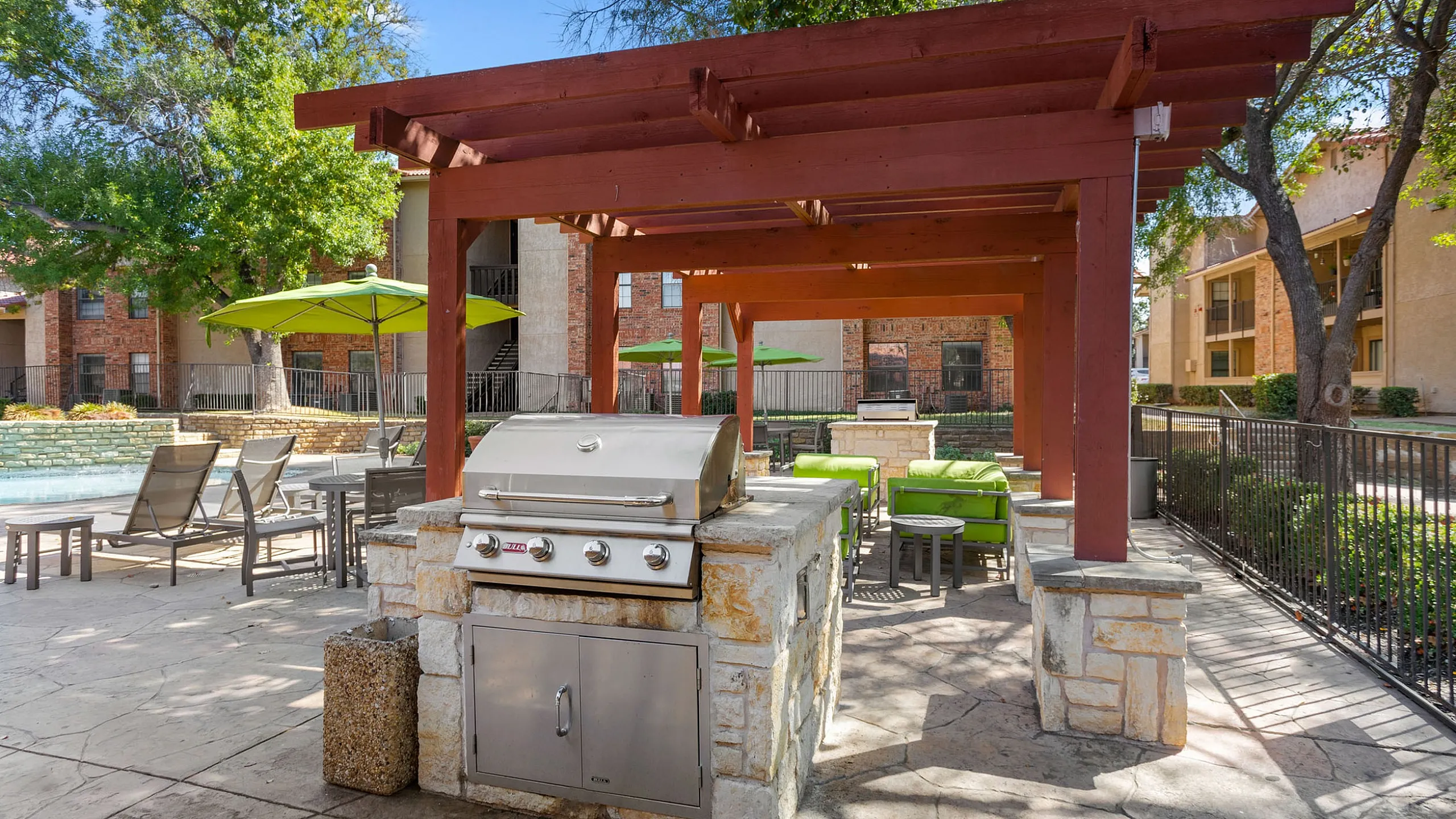 Inviting outdoor patio area with a modern grill, shaded by a wooden pergola, surrounded by green seating and vibrant umbrellas in a serene apartment community.