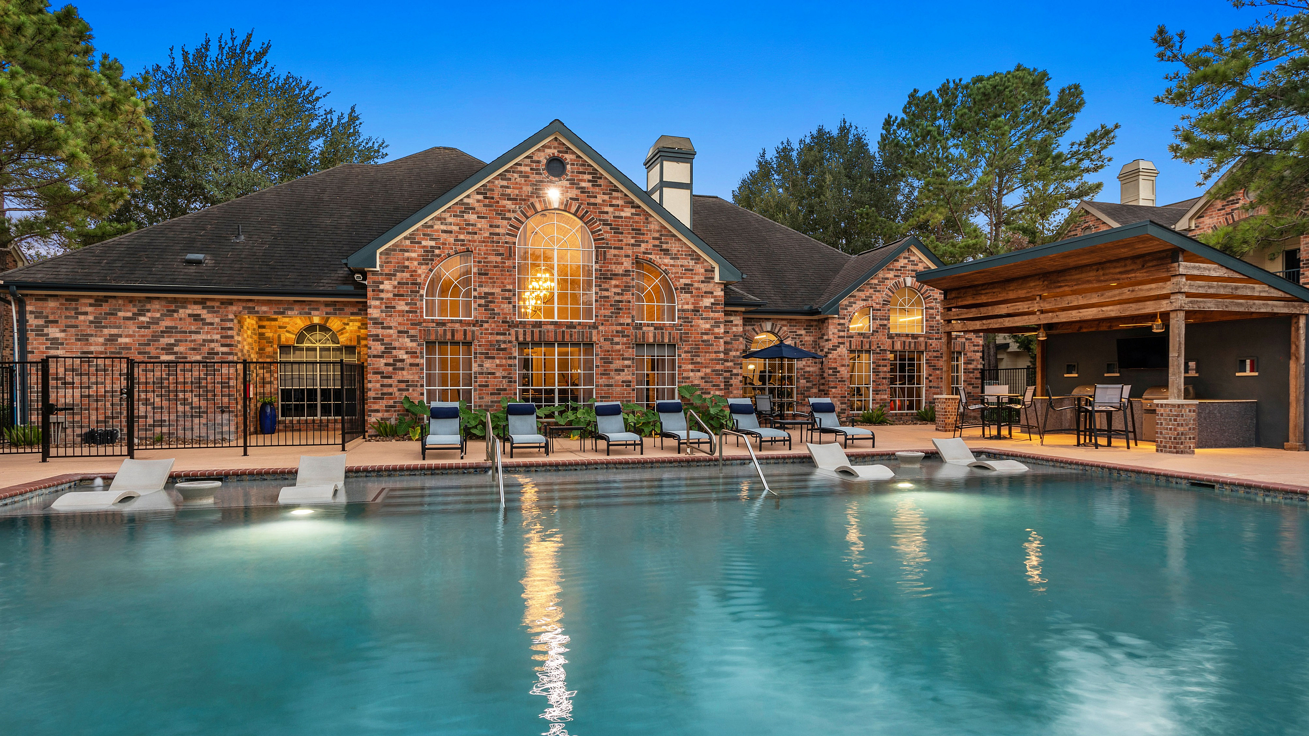 Charming brick apartment complex with expansive pool, lounge chairs, and outdoor seating area at dusk.