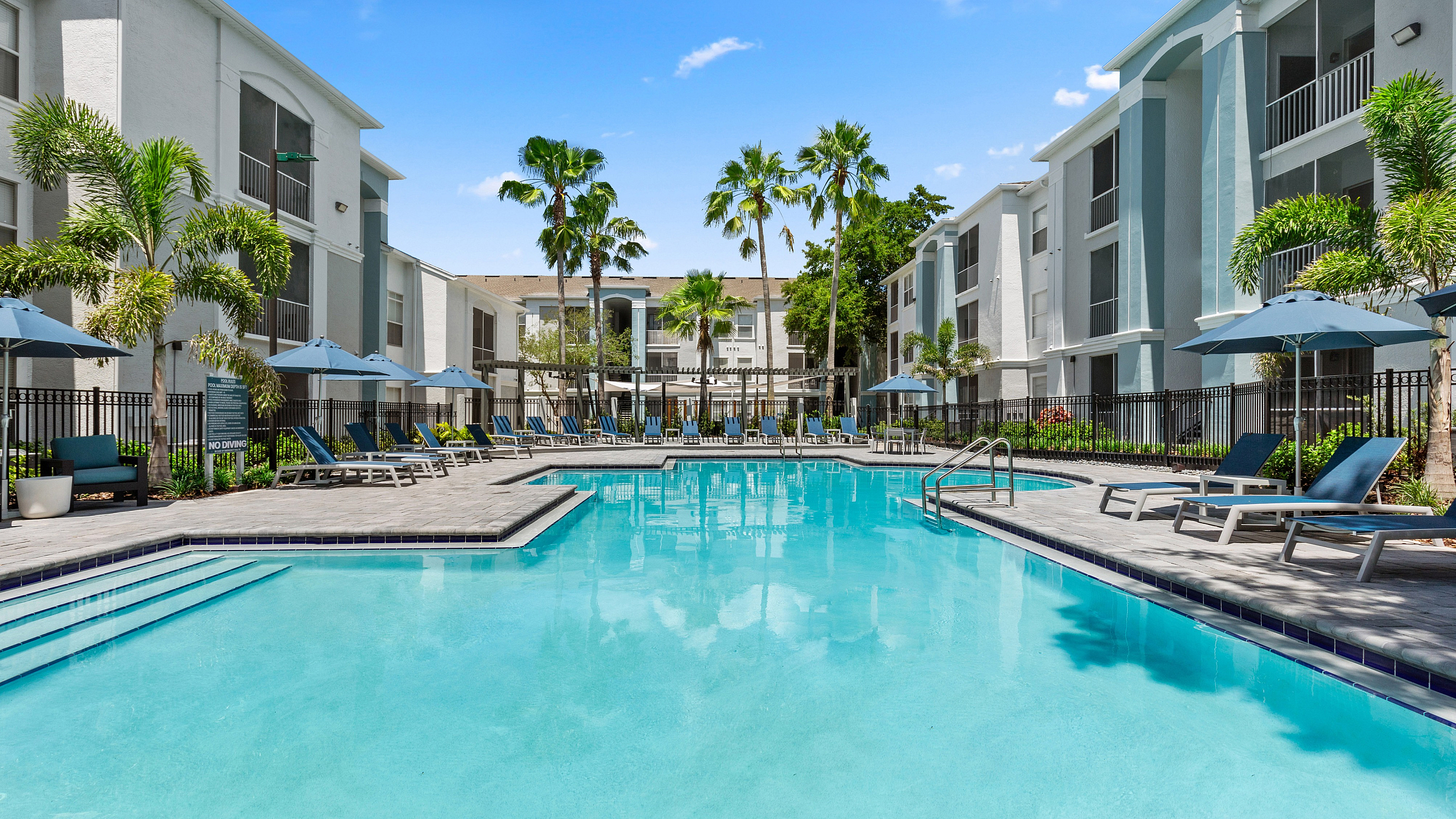Luxurious apartment complex with a pristine swimming pool, surrounded by palm trees and inviting lounge chairs under stylish umbrellas.
