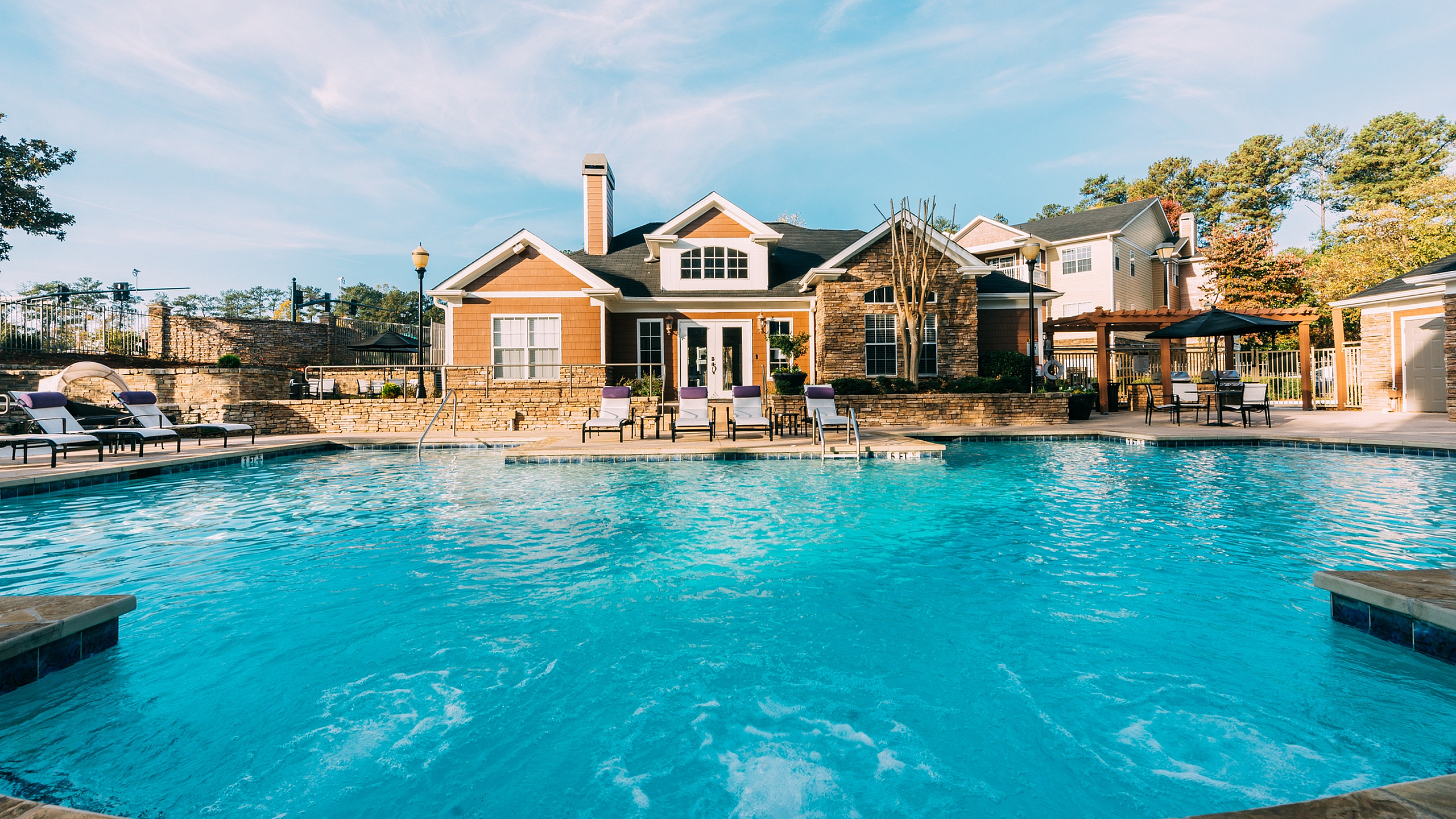 Luxurious apartment complex with a beautiful sparkling pool, inviting lounge chairs, and elegant stone-accented architecture under a clear blue sky.