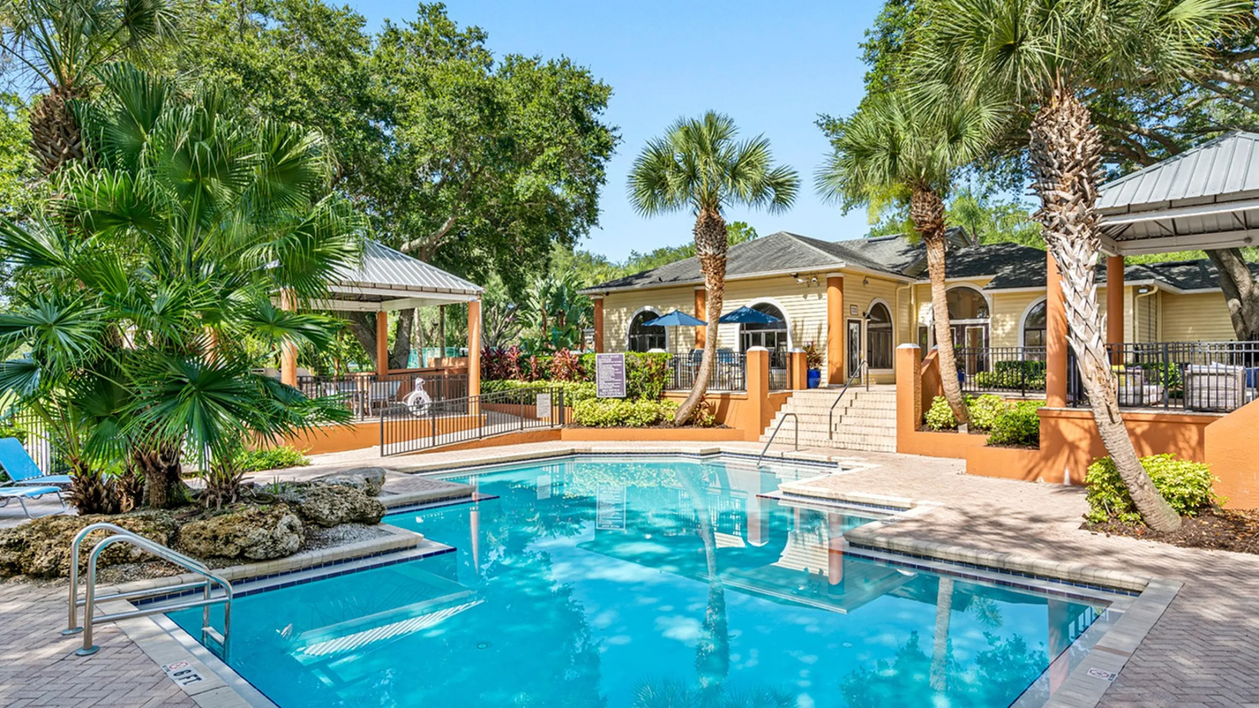 Charming outdoor pool area with lush palm trees, shaded seating, and inviting clubhouse.