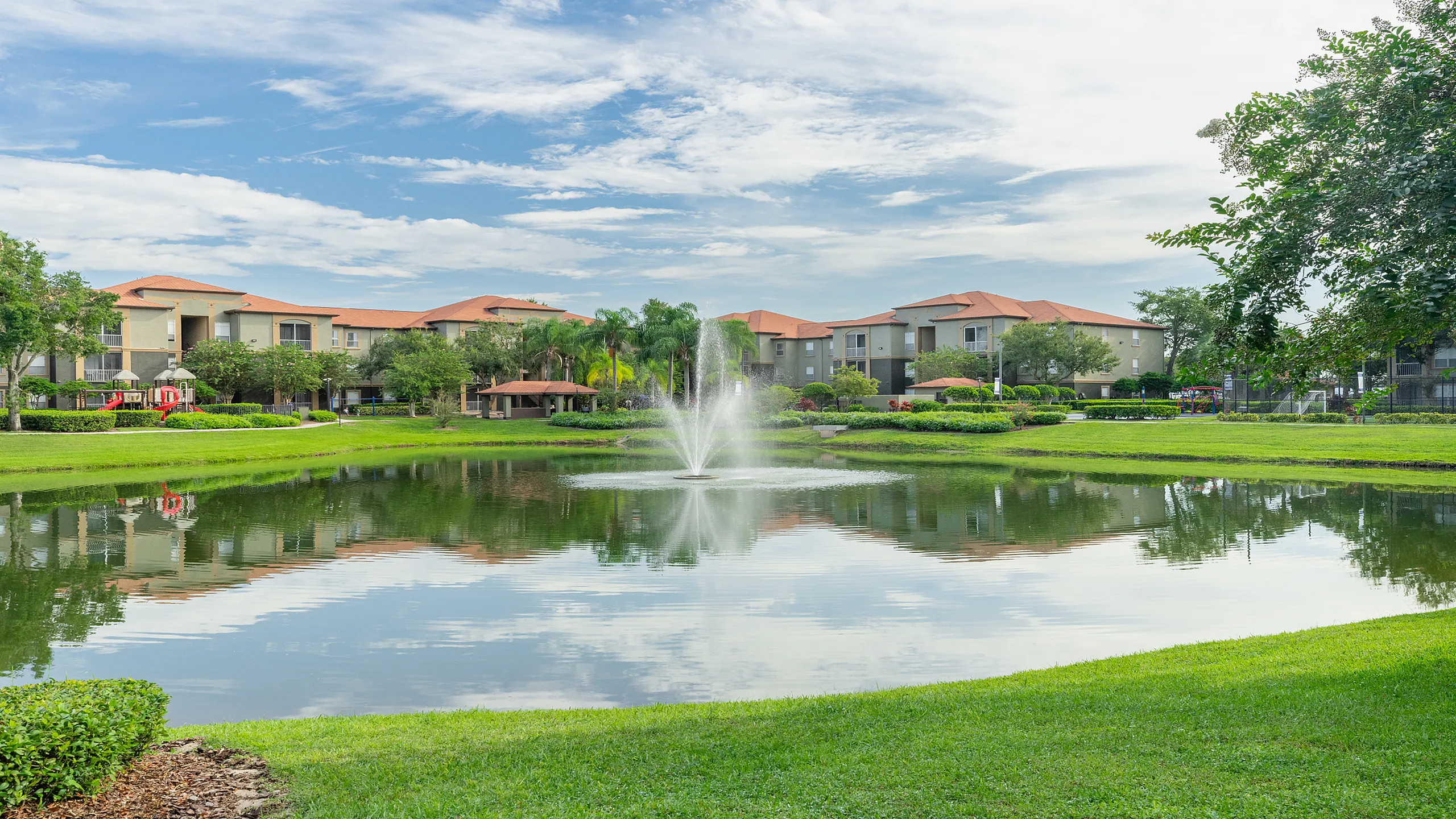 Charming apartment complex with a serene pond, central fountain, and lush greenery, offering tranquil waterside living.