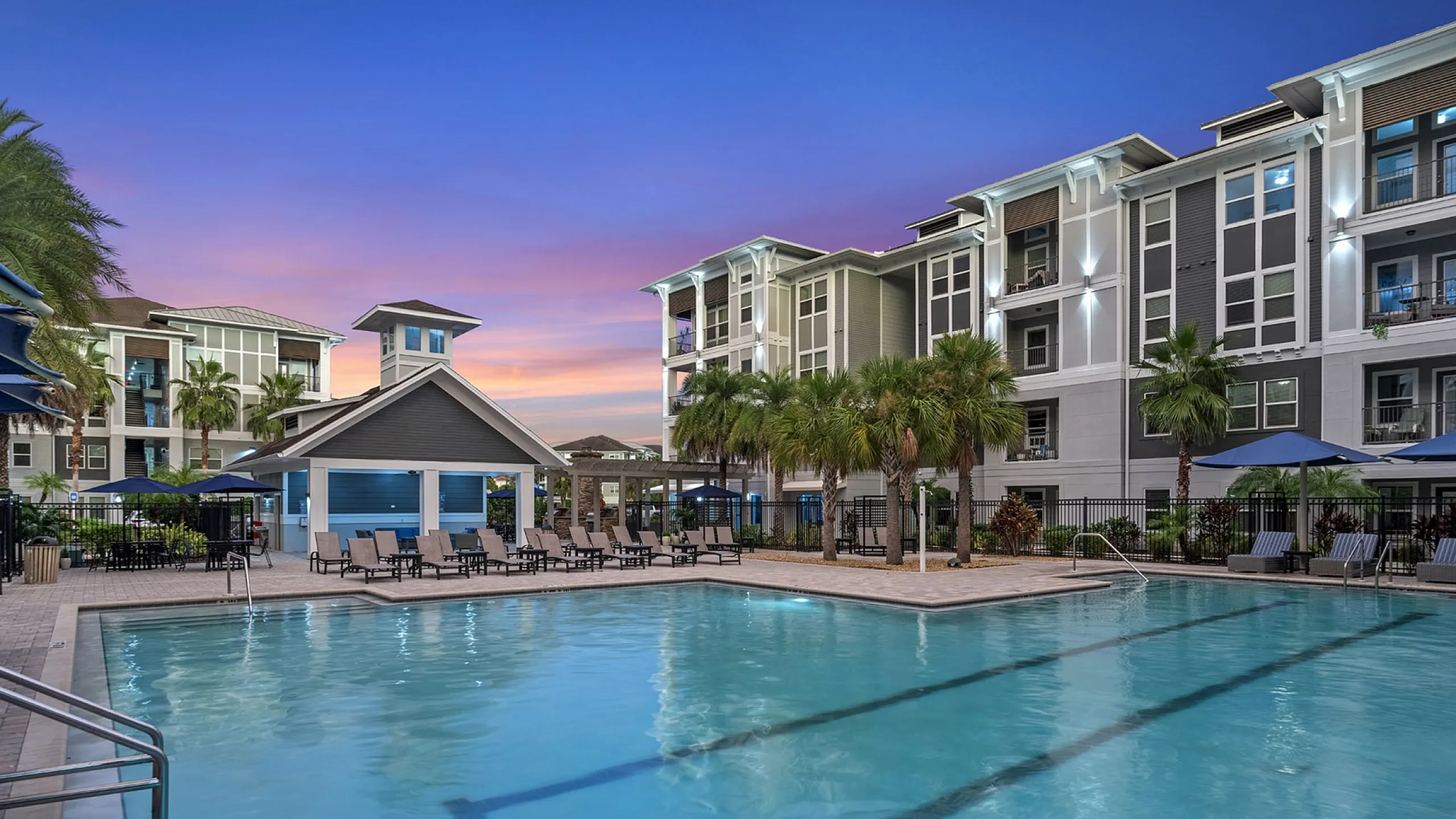 Exquisite apartment complex with a shimmering pool, surrounded by elegant palm trees and stylish seating, set against a vibrant evening sky.