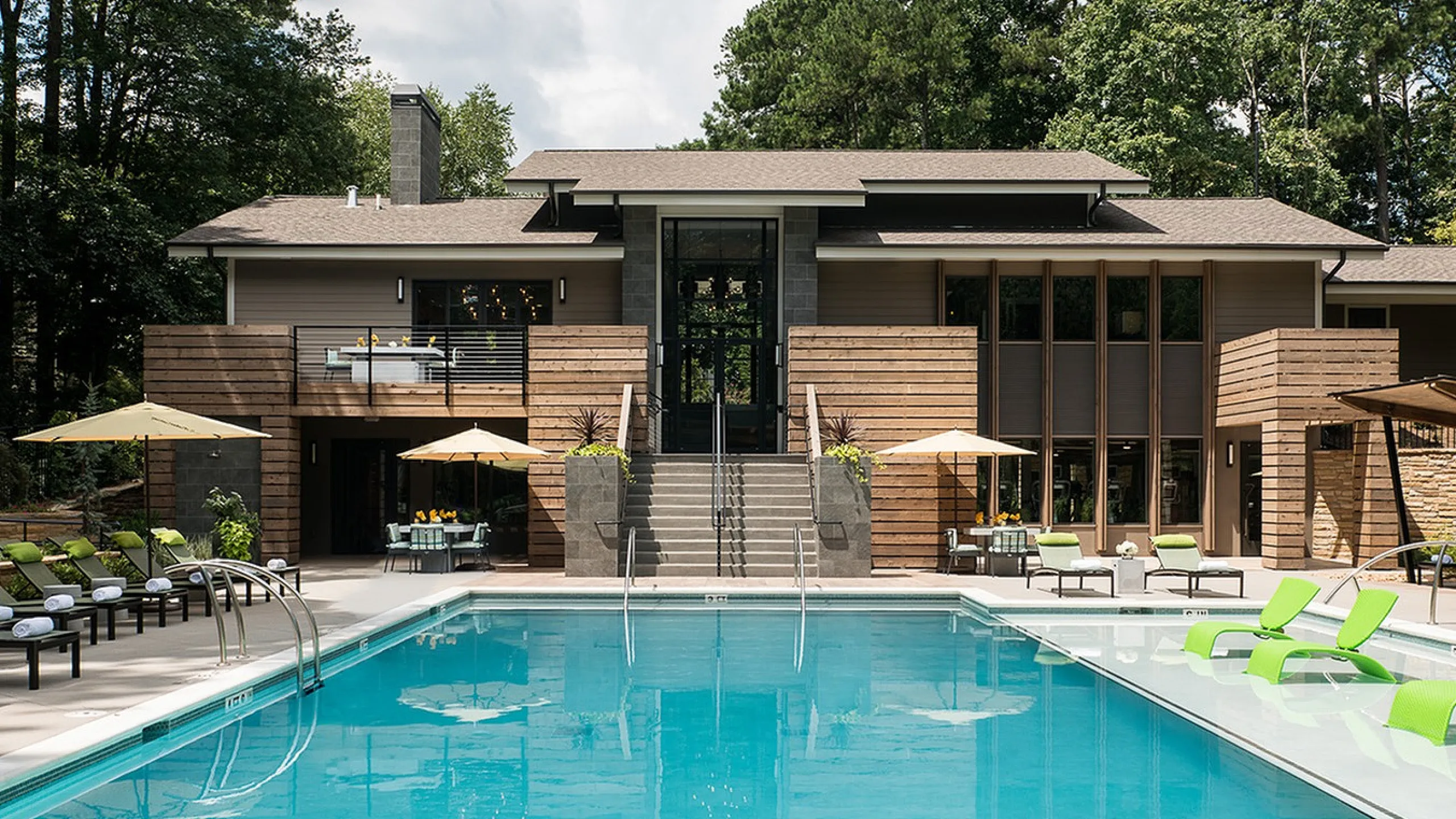 Elegant apartment exterior featuring modern wood paneling, a luxurious swimming pool with vibrant green lounge chairs, and stylish outdoor seating with umbrellas, set against a lush forest backdrop.