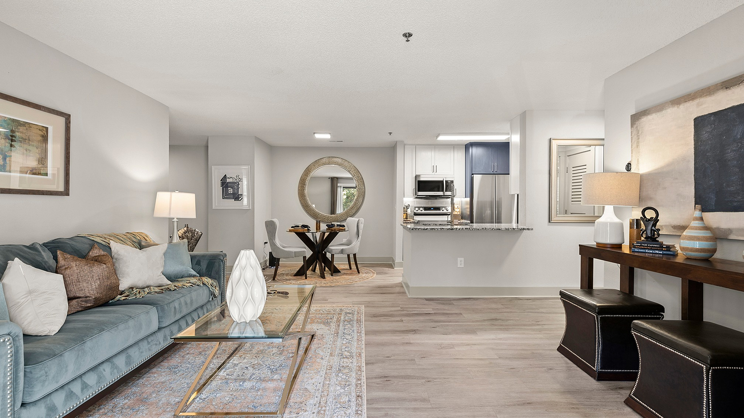 Inviting open-concept living space with elegant modern furnishings, featuring a plush blue sofa, glass coffee table, and contemporary art. Seamlessly connected dining area and sleek kitchen with stainless steel appliances.