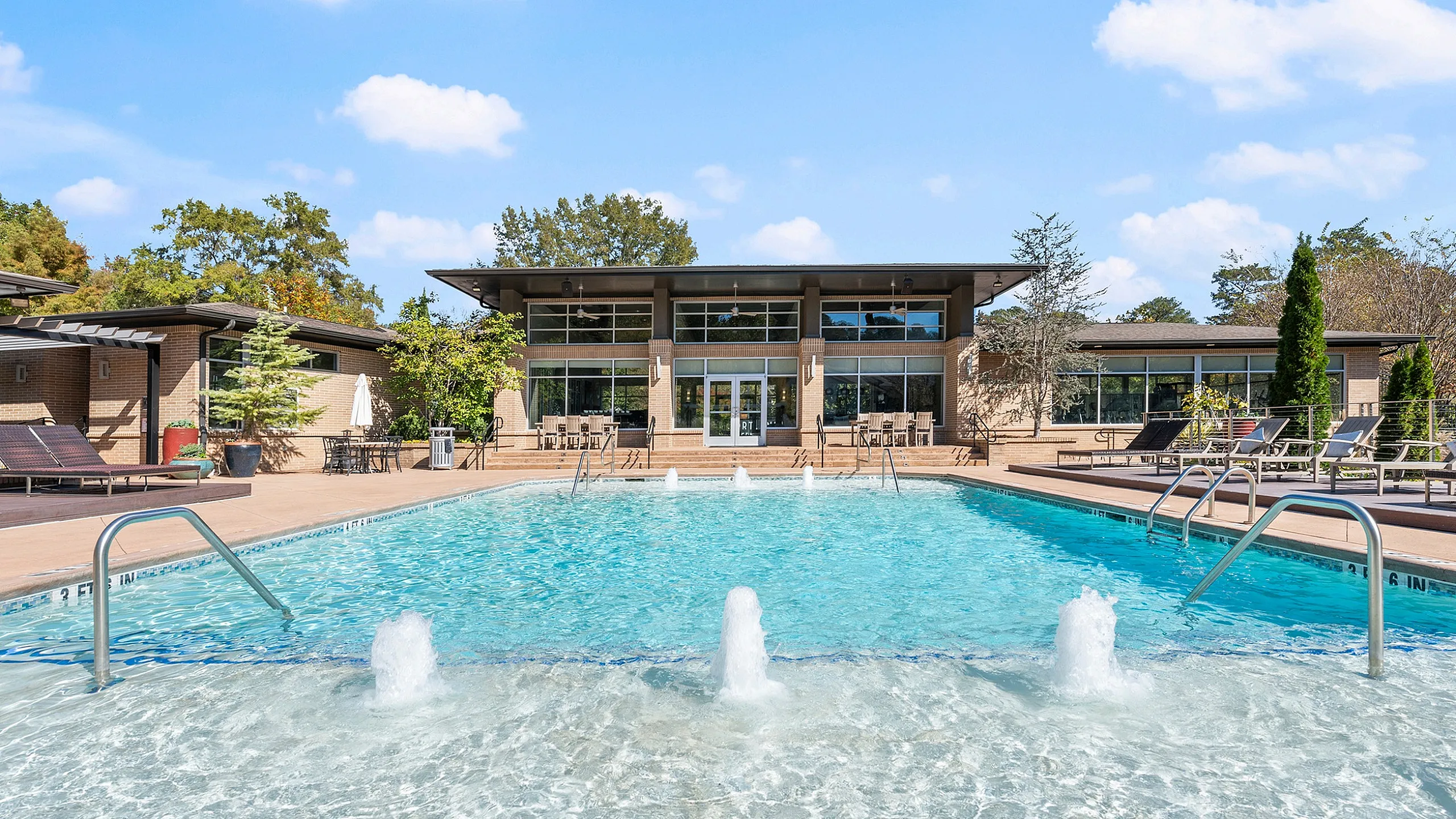 Luxurious outdoor swimming pool with water features, surrounded by sun loungers and a modern clubhouse.