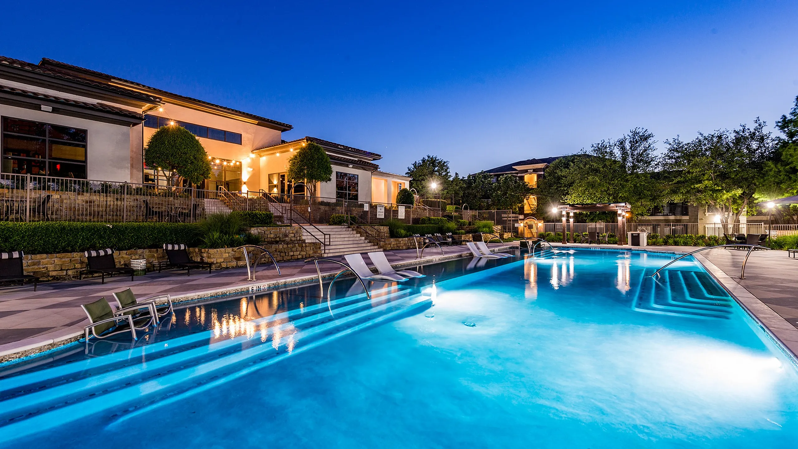 Luxurious outdoor pool area with stylish lounge chairs, elegant landscaping, and modern architecture illuminated under a twilight sky.