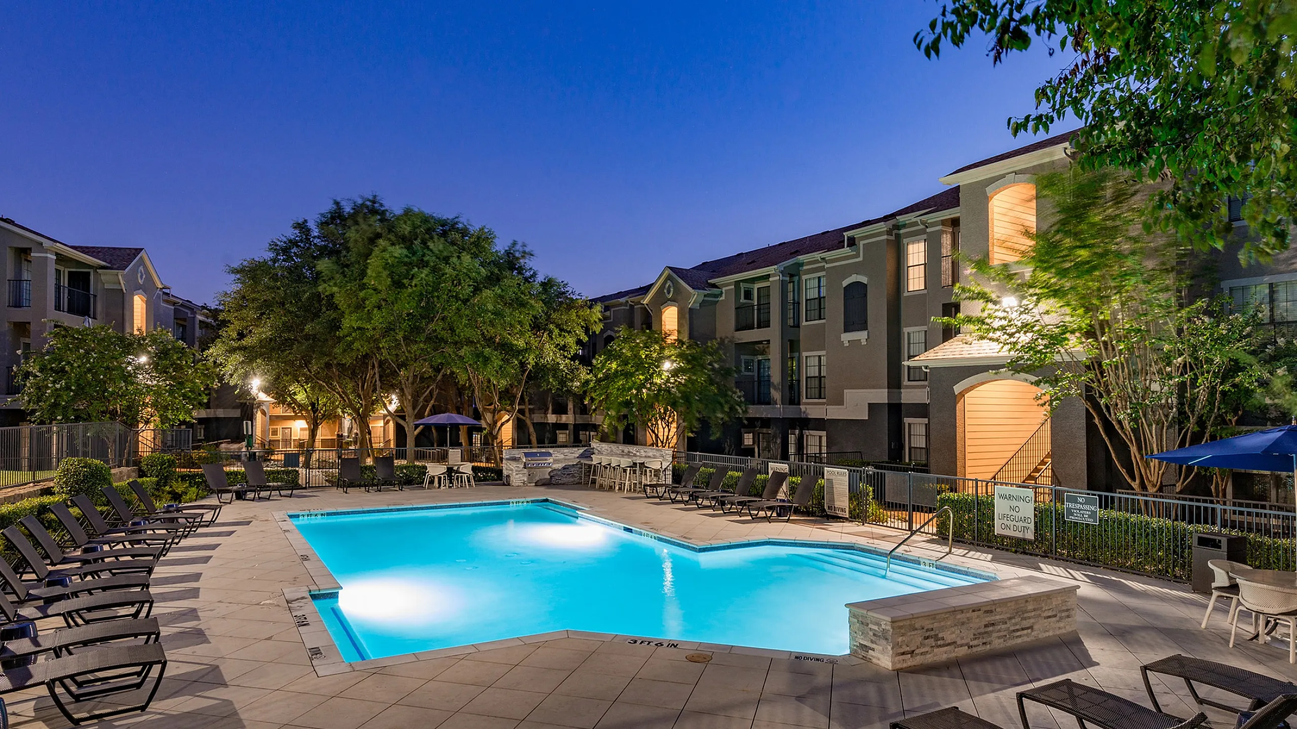 Elegant apartment complex with a beautifully lit outdoor pool area surrounded by lush trees and modern buildings at dusk.
