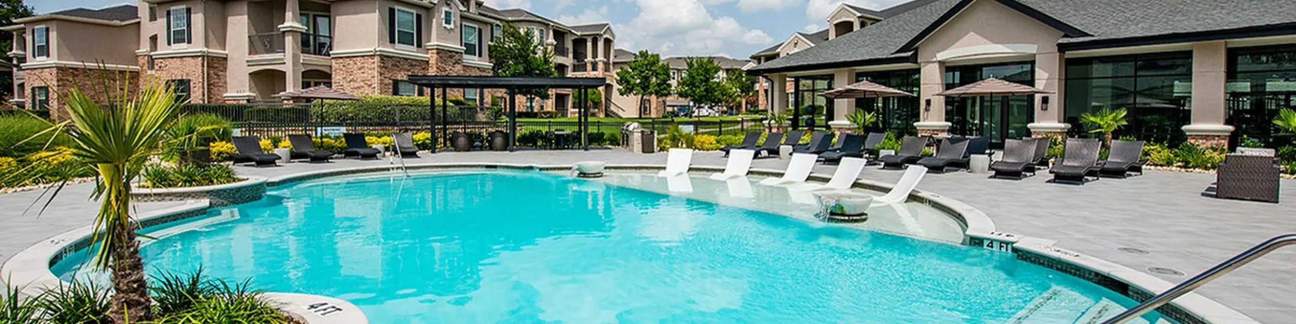 Luxurious apartment complex featuring a stunning pool area with sun loungers, lush landscaping, and beautifully designed buildings under a clear blue sky.