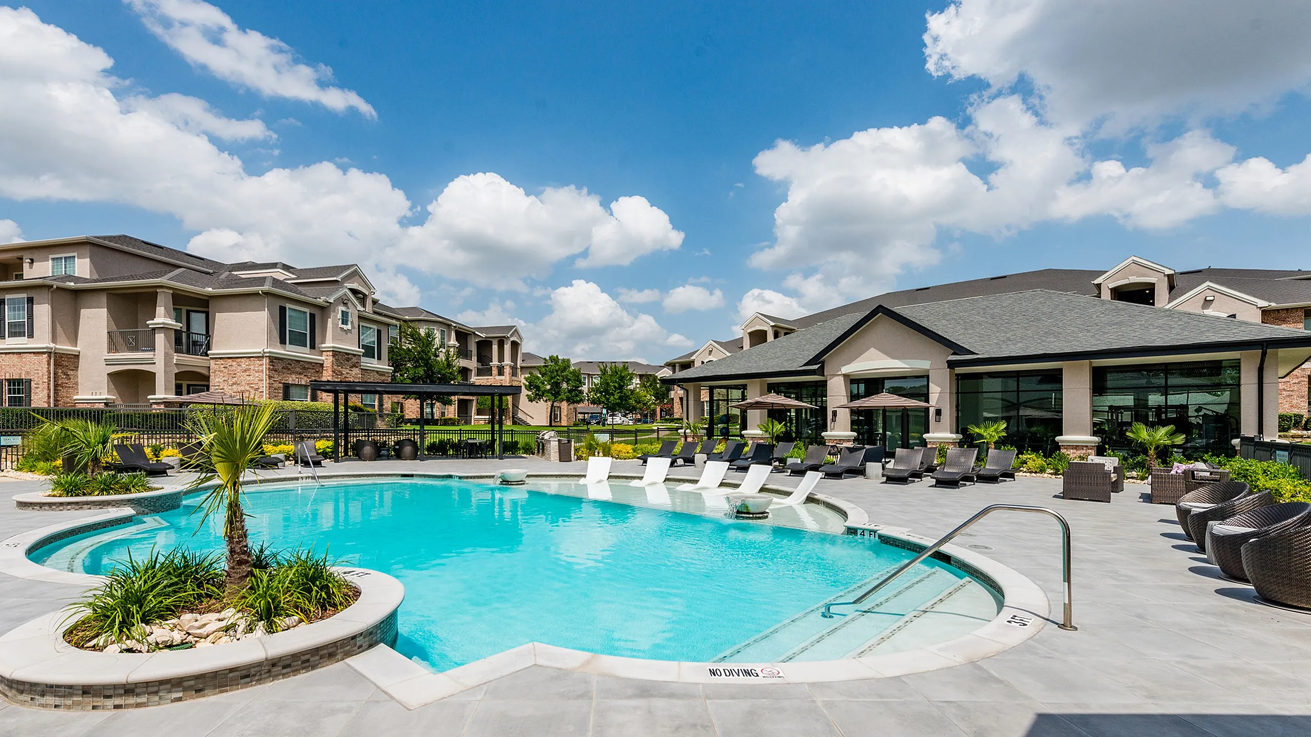 Luxurious apartment complex with a pristine swimming pool, stylish lounge chairs, and beautifully landscaped surroundings under a vibrant blue sky.