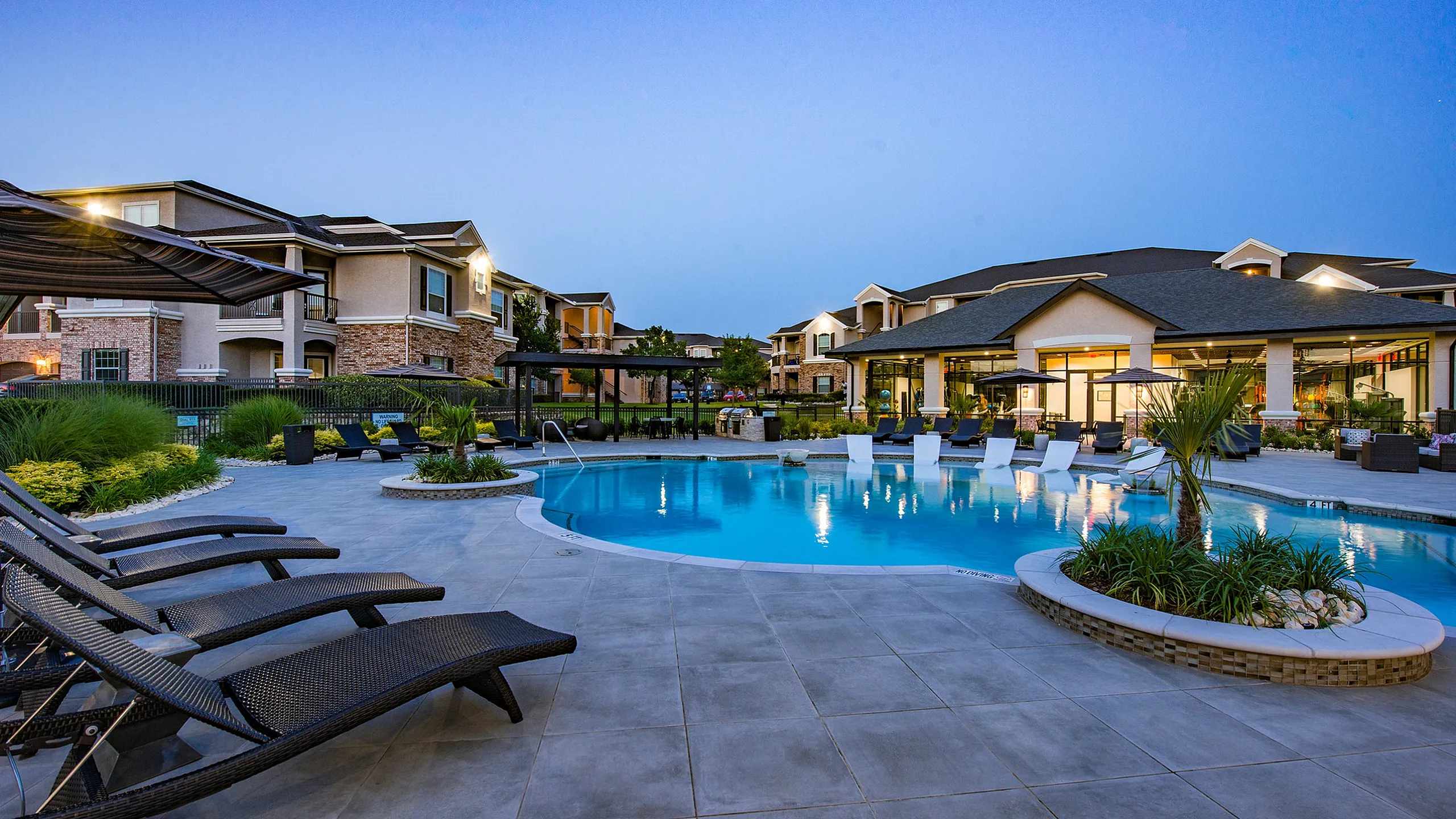 Luxurious apartment complex with an inviting poolside setting, featuring elegant sun loungers and serene landscaping under a stunning evening sky.