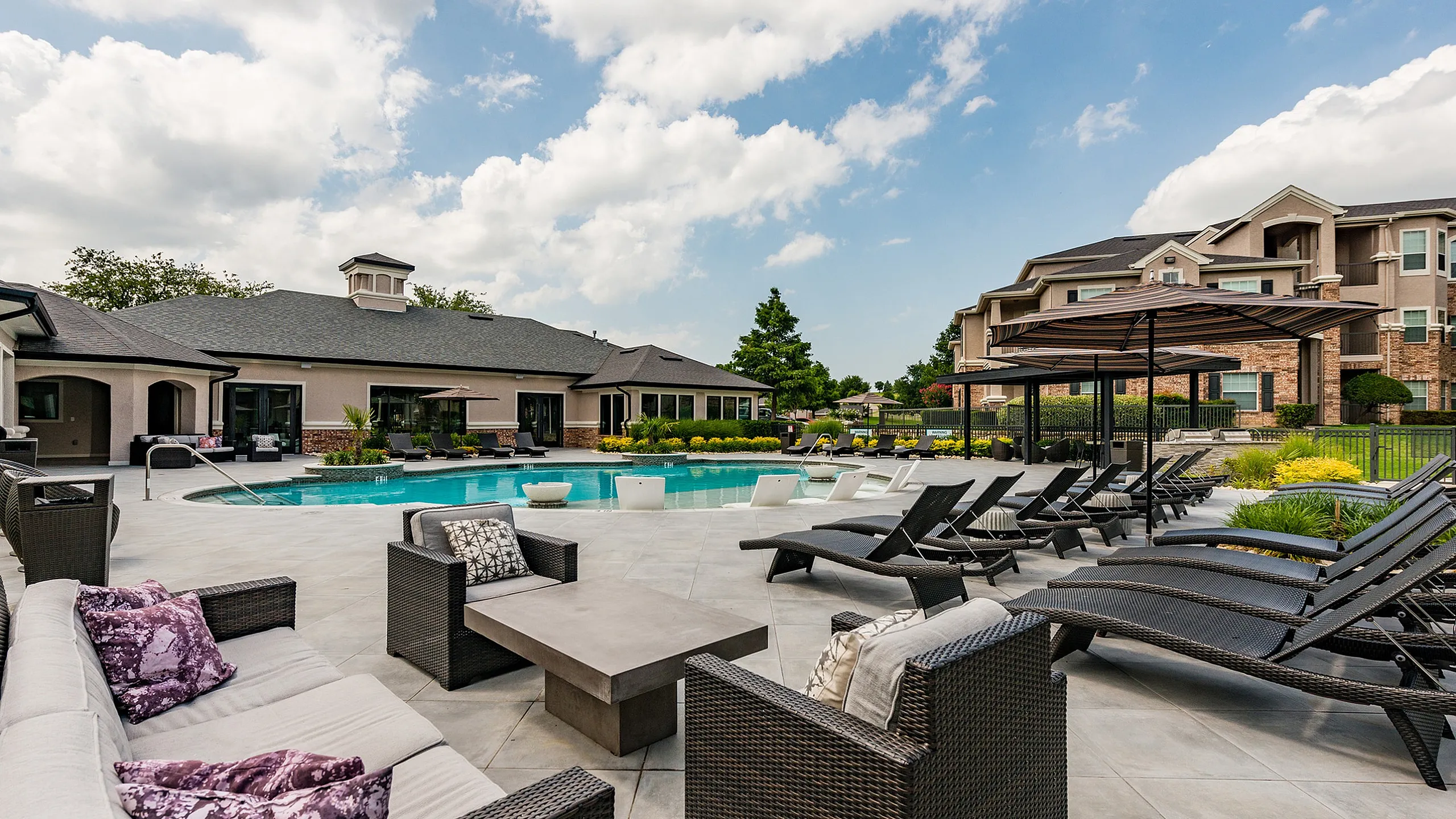 Luxurious outdoor pool area featuring elegant lounge chairs, cozy seating, and vibrant landscaping against a sunny sky backdrop.