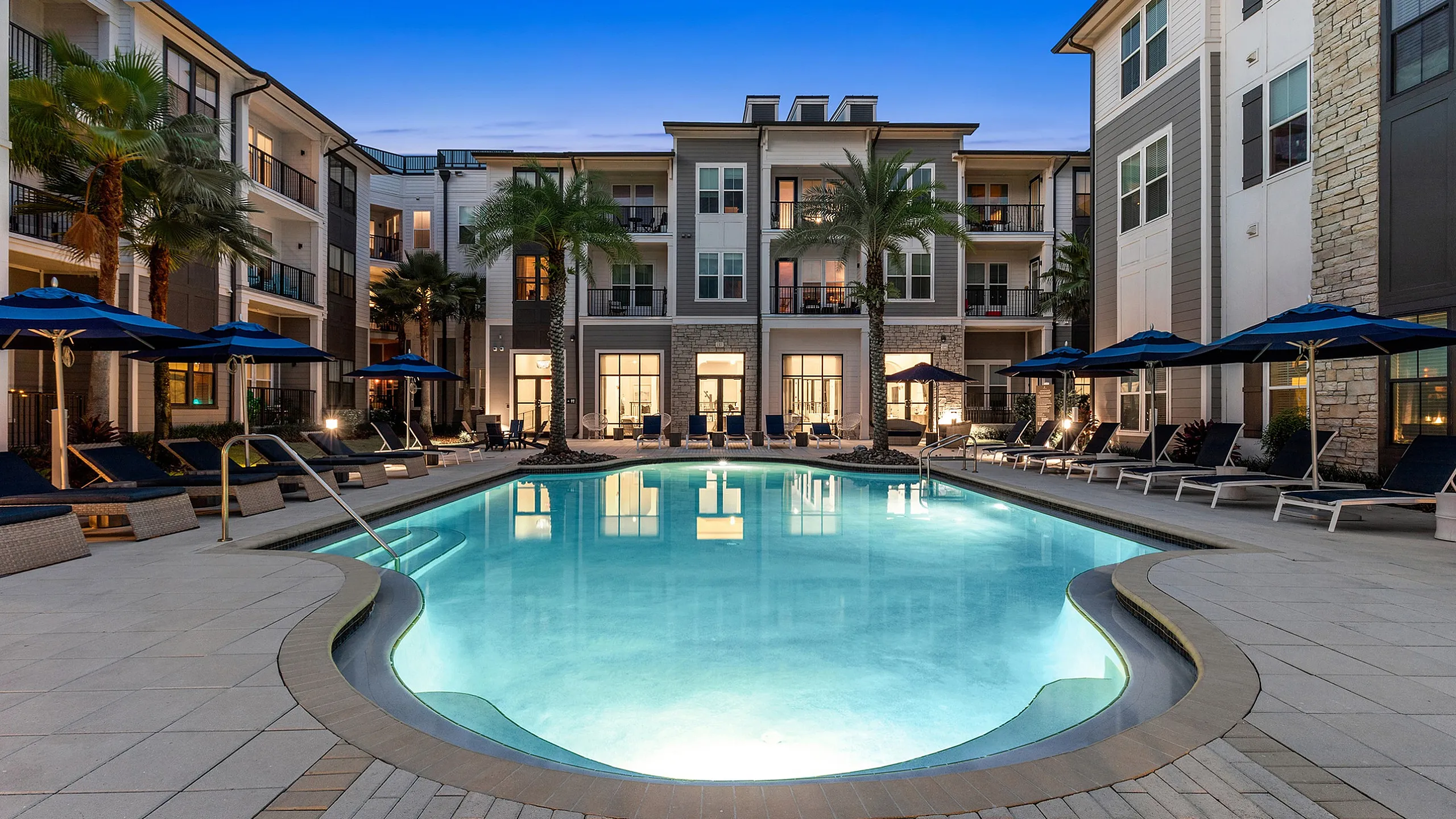 Luxurious apartment complex featuring a glowing pool with elegant lounge chairs, surrounded by modern architecture and palm trees at dusk.