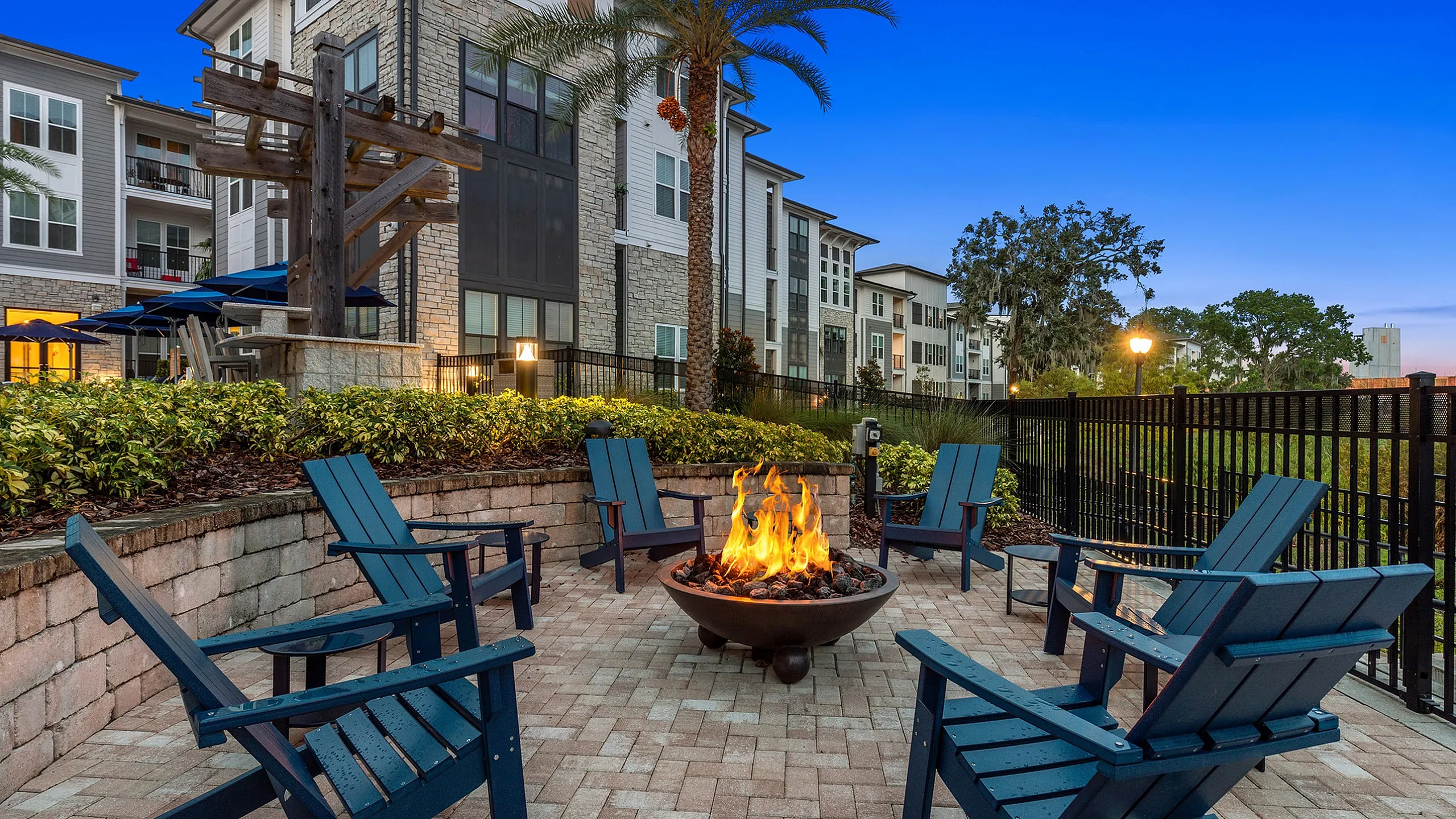 Charming outdoor seating area with Adirondack chairs surrounding a cozy fire pit, framed by lush greenery and elegant apartment balconies under a twilight sky.