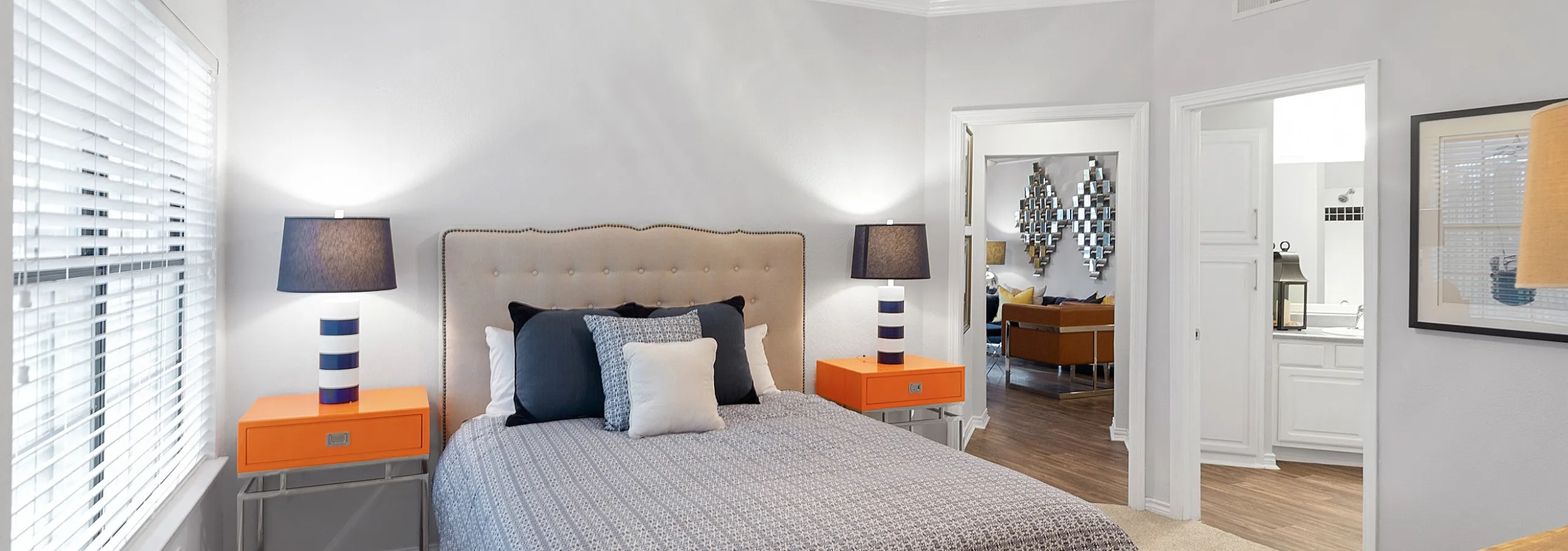Elegant bedroom with modern orange nightstands, luxurious bedding, and ceiling fan, leading to a chic living area in a stylish apartment.