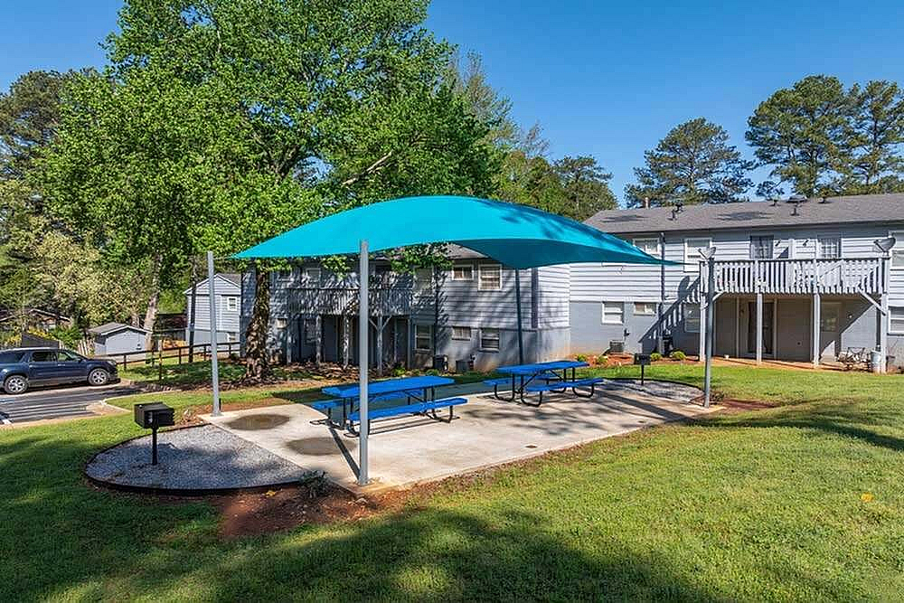 Charming outdoor space with vibrant blue picnic tables under a shade sail, perfect for gatherings in a lush green setting.