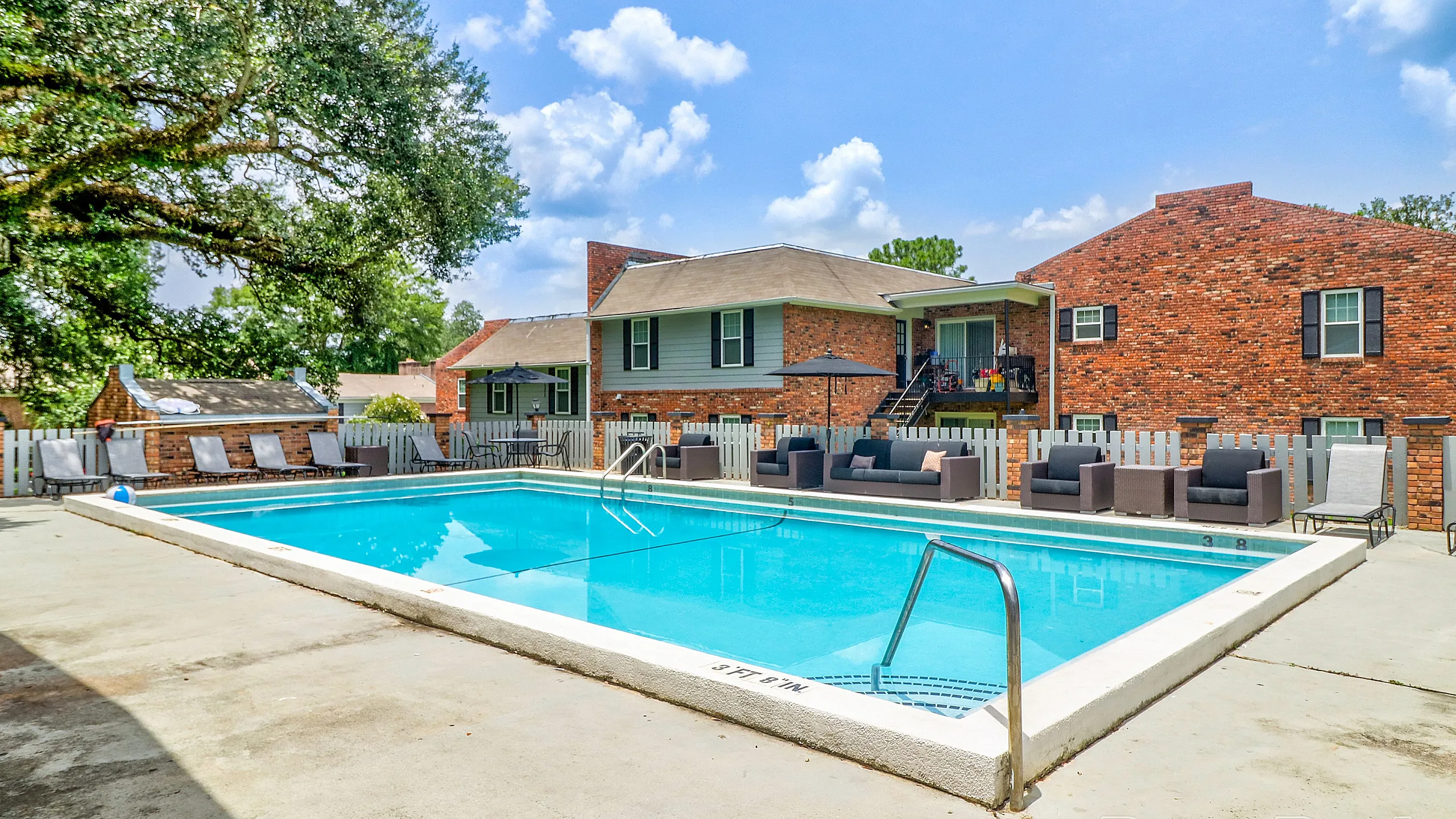Inviting outdoor pool area with comfortable seating, set against a charming brick apartment backdrop and lush greenery.