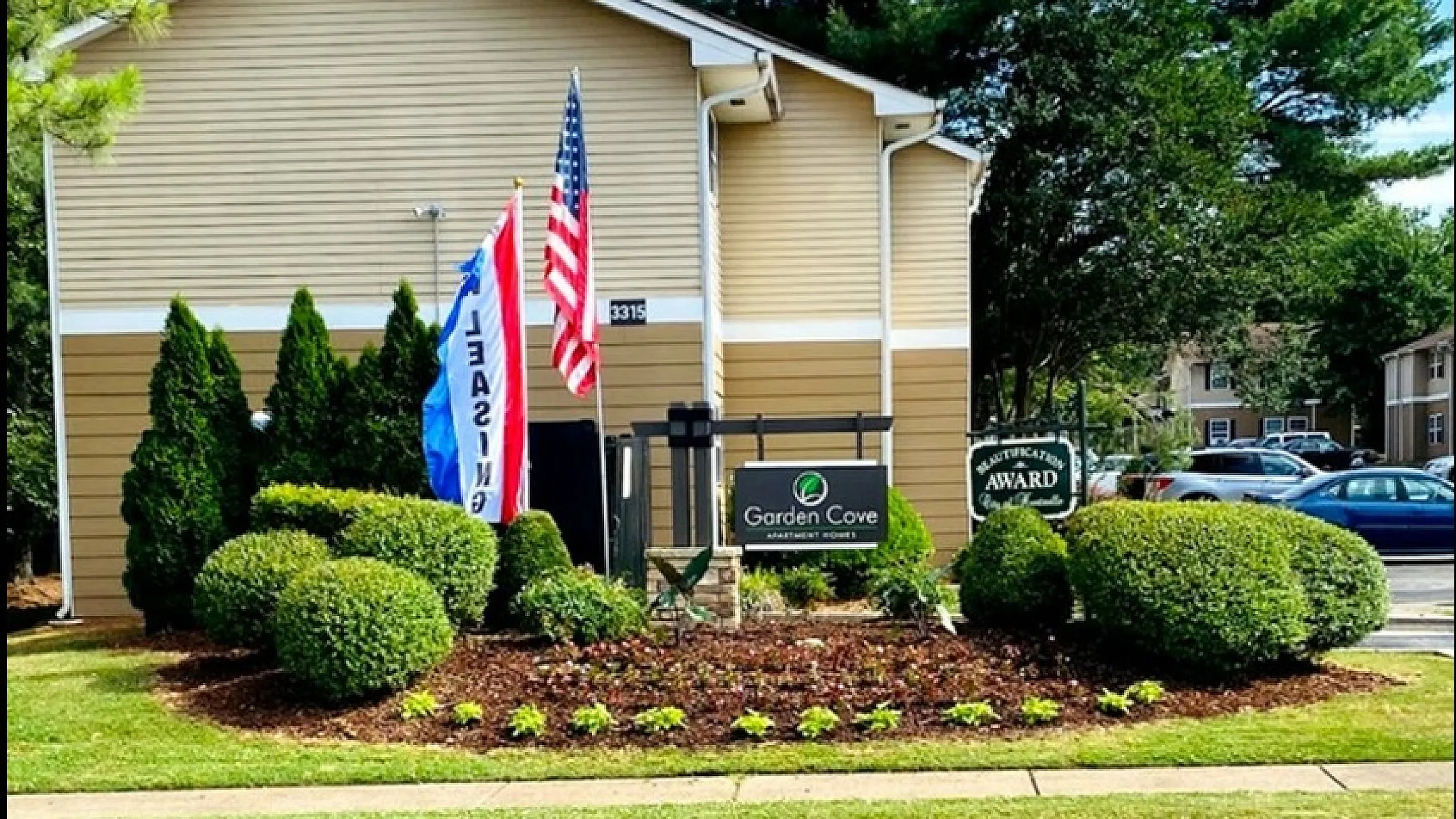 Charming Garden Cove Apartments entrance surrounded by lush greenery and vibrant flags, showcasing welcoming community vibes.