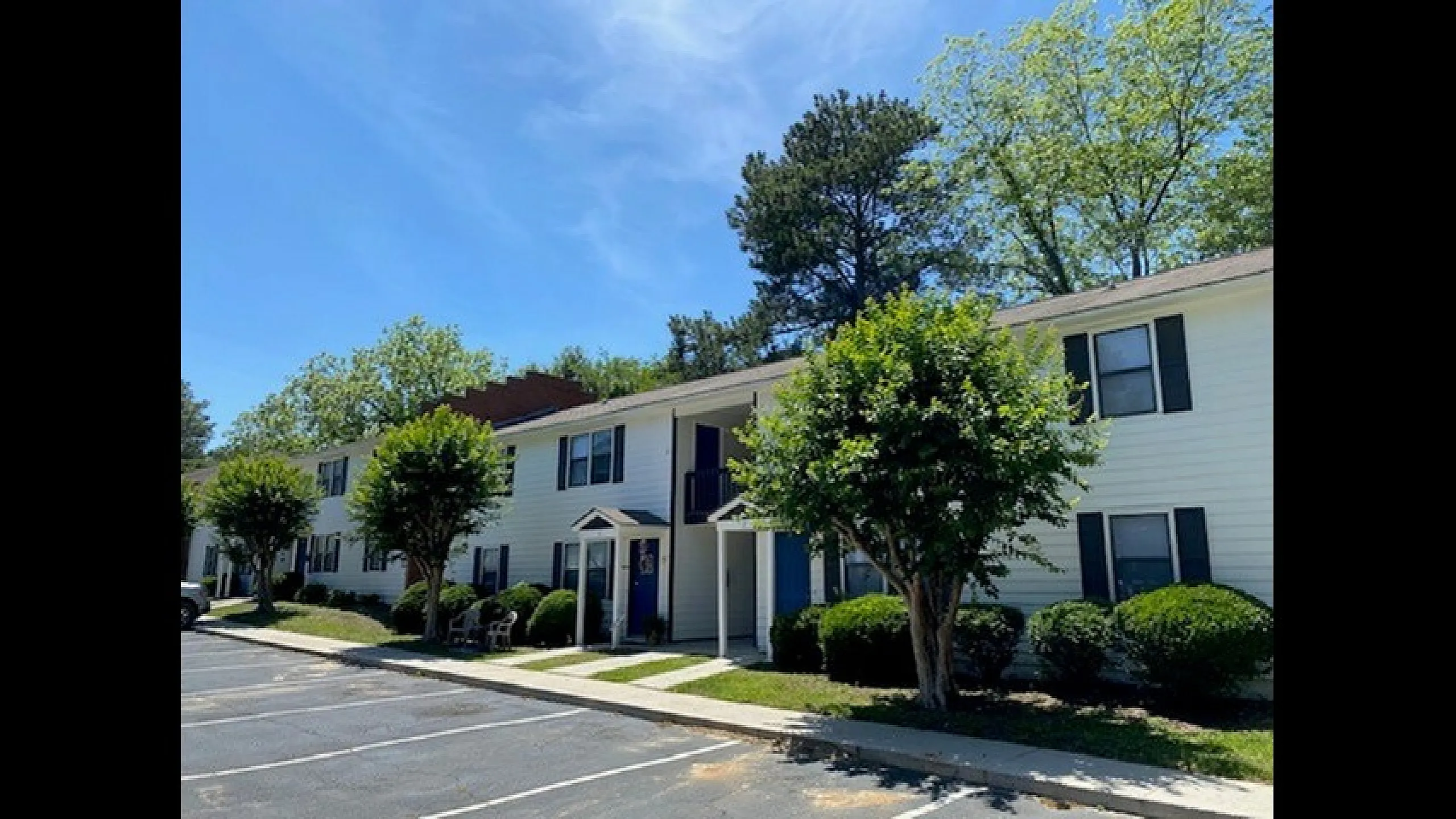 Charming two-story apartment building with white siding, surrounded by lush greenery and mature trees under a clear blue sky.