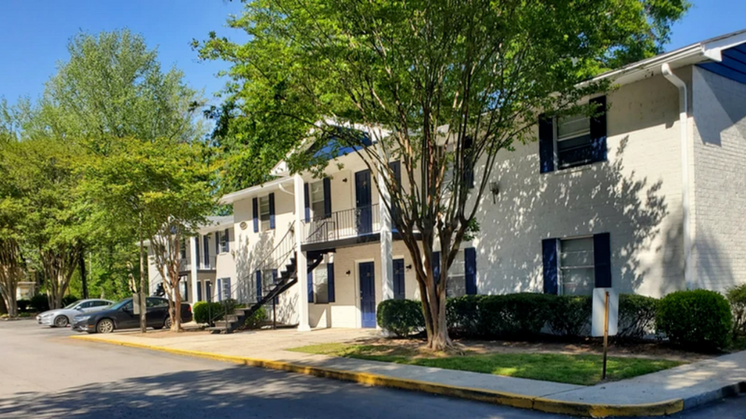 Charming apartment complex with white brick exterior, surrounded by lush greenery and ample parking.