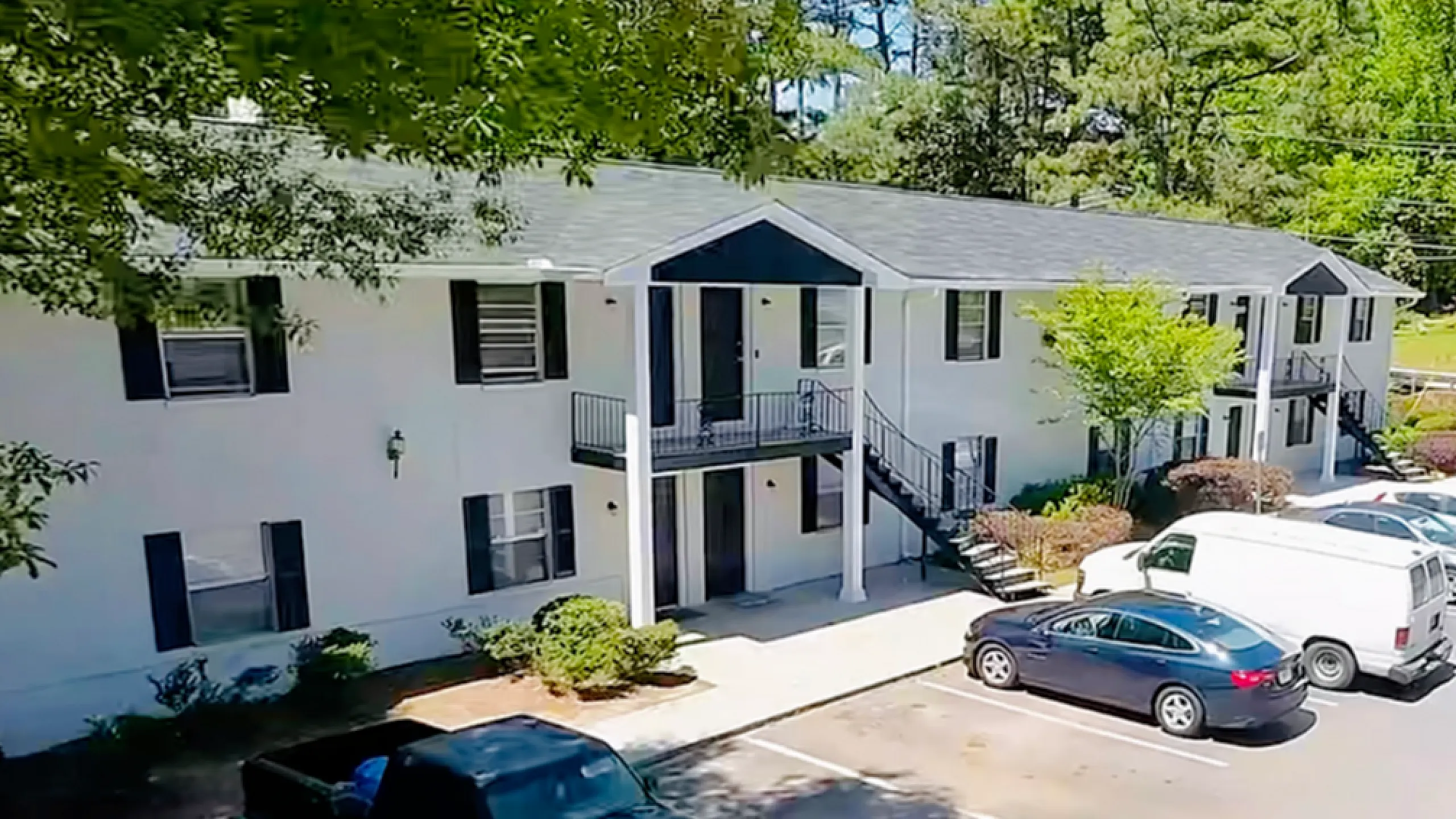 Charming white two-story apartment complex with black shutters, nestled among lush trees, featuring convenient parking and modern outdoor staircases.
